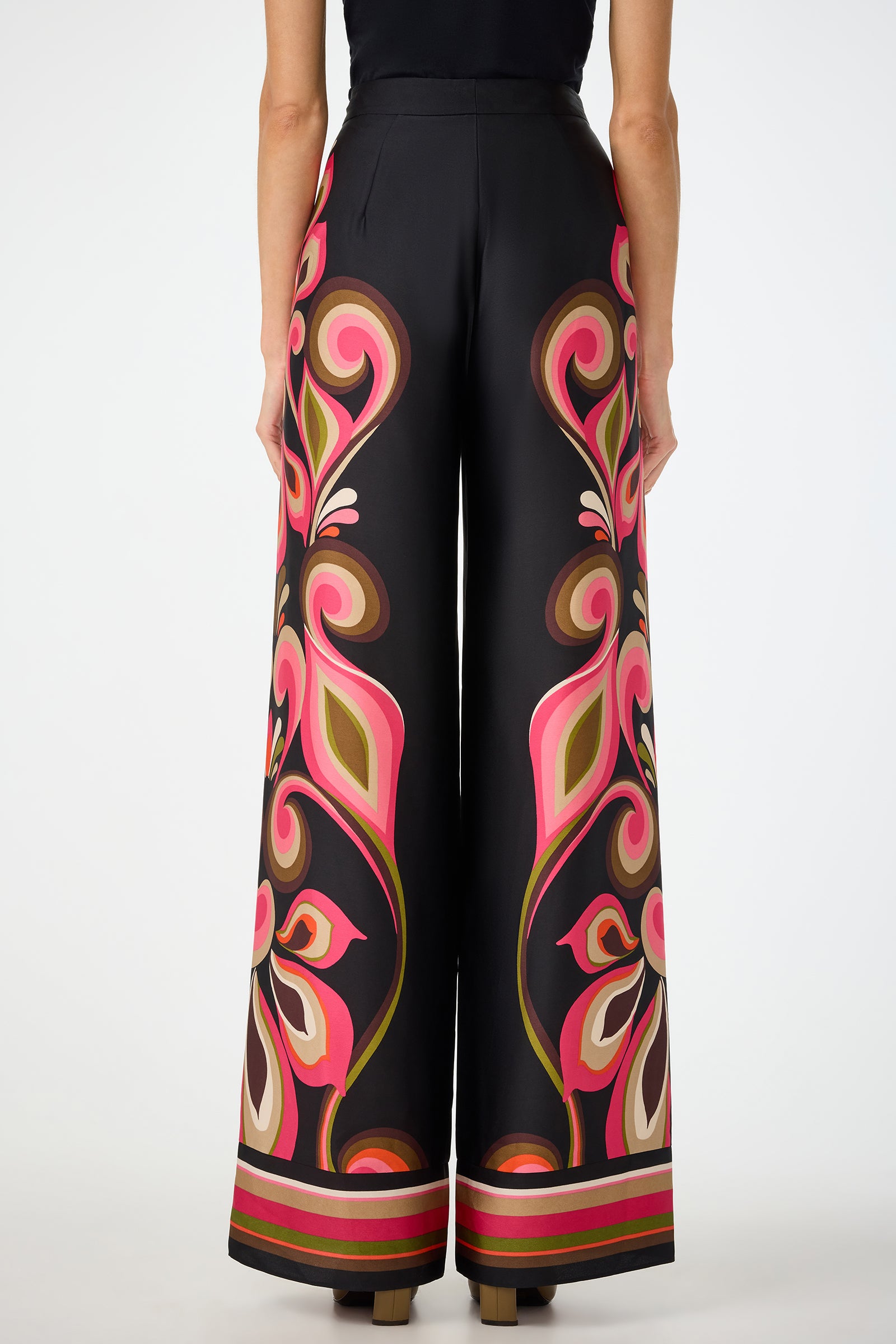A woman is seen from the back against a plain background wearing Verity Border Pants—high-waisted, wide-leg black pants featuring bold floral and swirl patterns in pink, brown, olive, and beige.