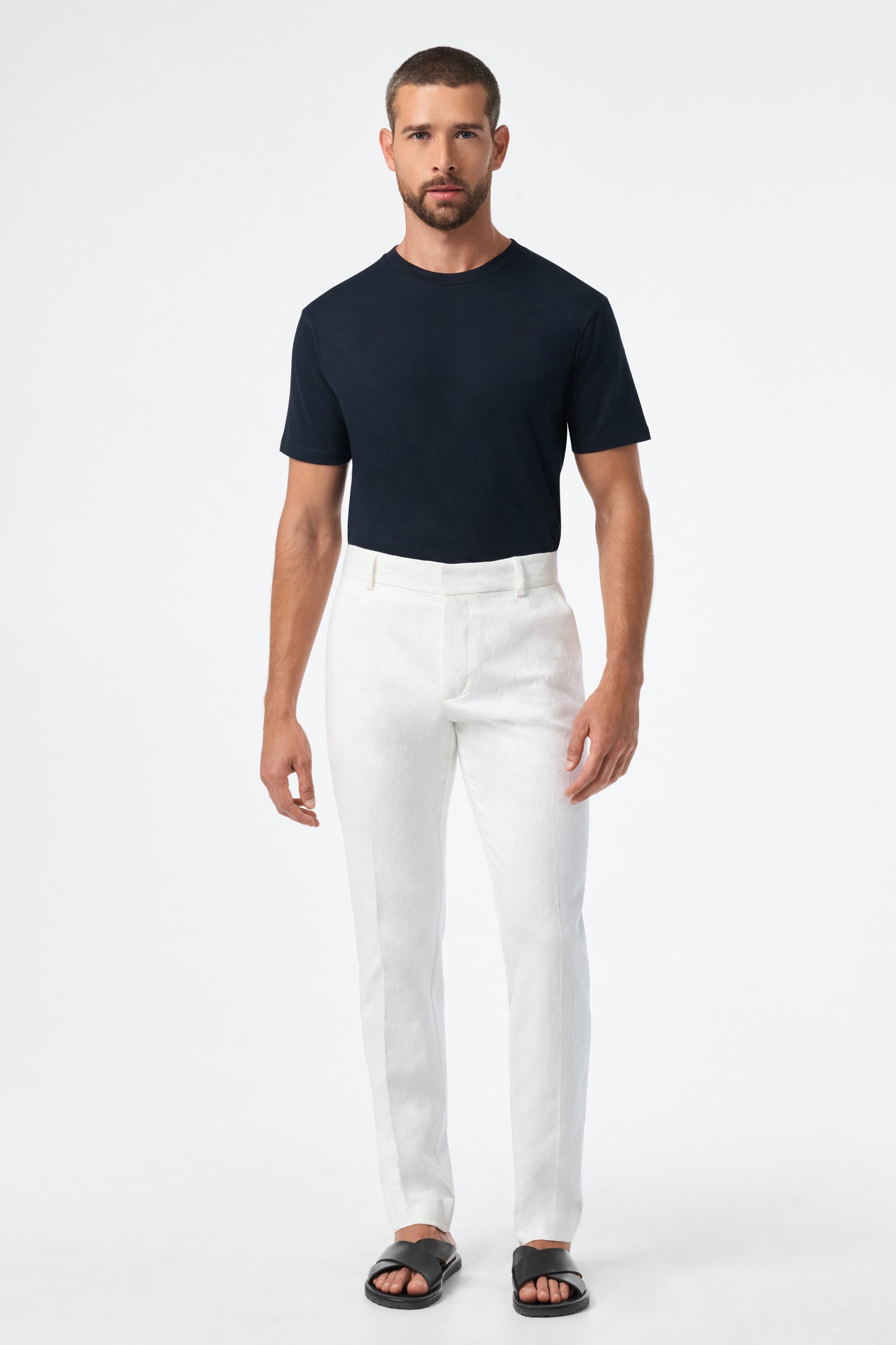 A man stands facing forward against a plain white background, wearing a fitted black t-shirt, the Clyde Slim Trouser - Stretch Herringbone in white, and black sandals.