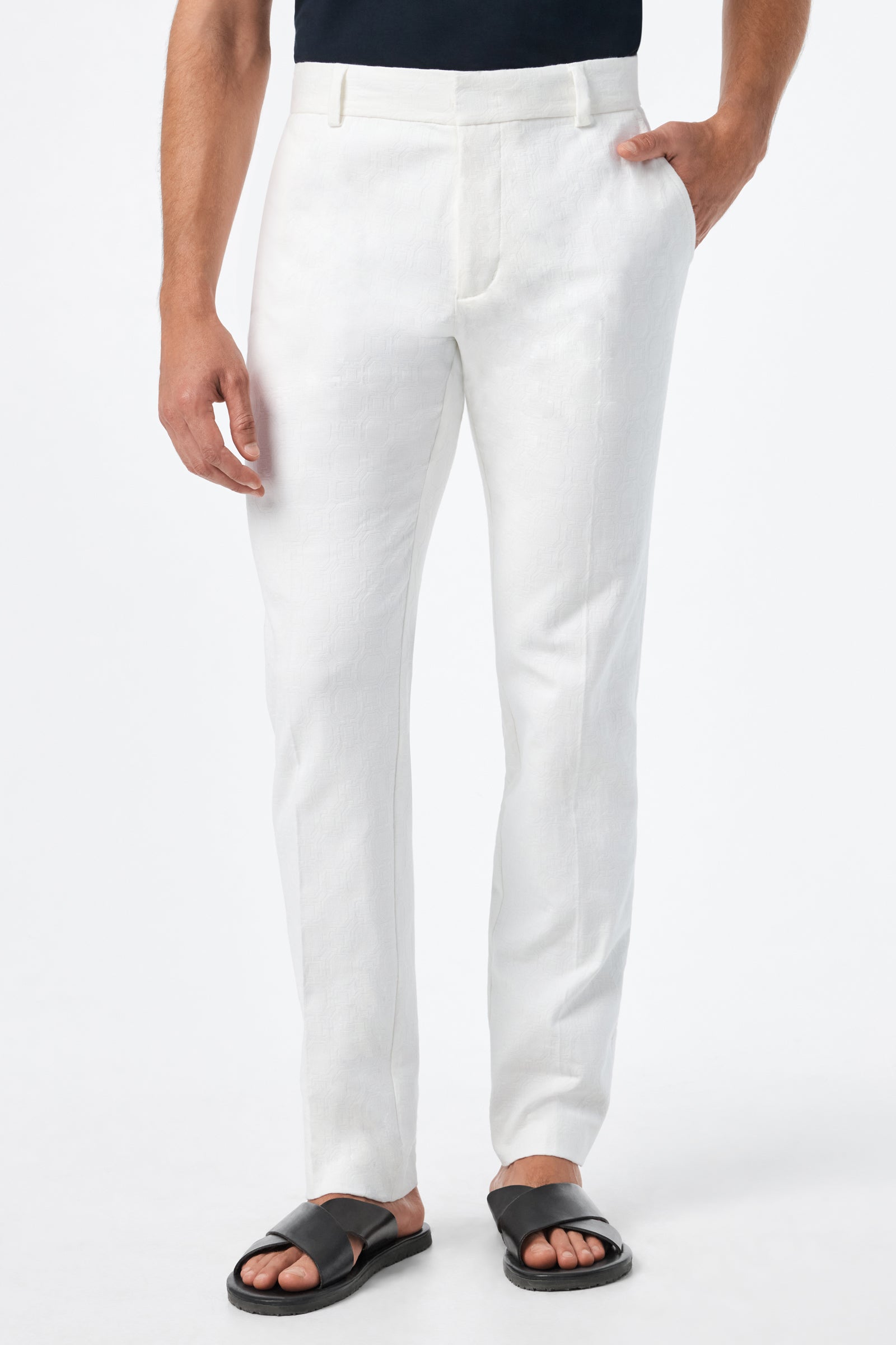 A person stands against a plain white background with one hand in their pocket, wearing the Clyde Slim Trouser - Stretch Herringbone, paired with a black shirt and black sandals.