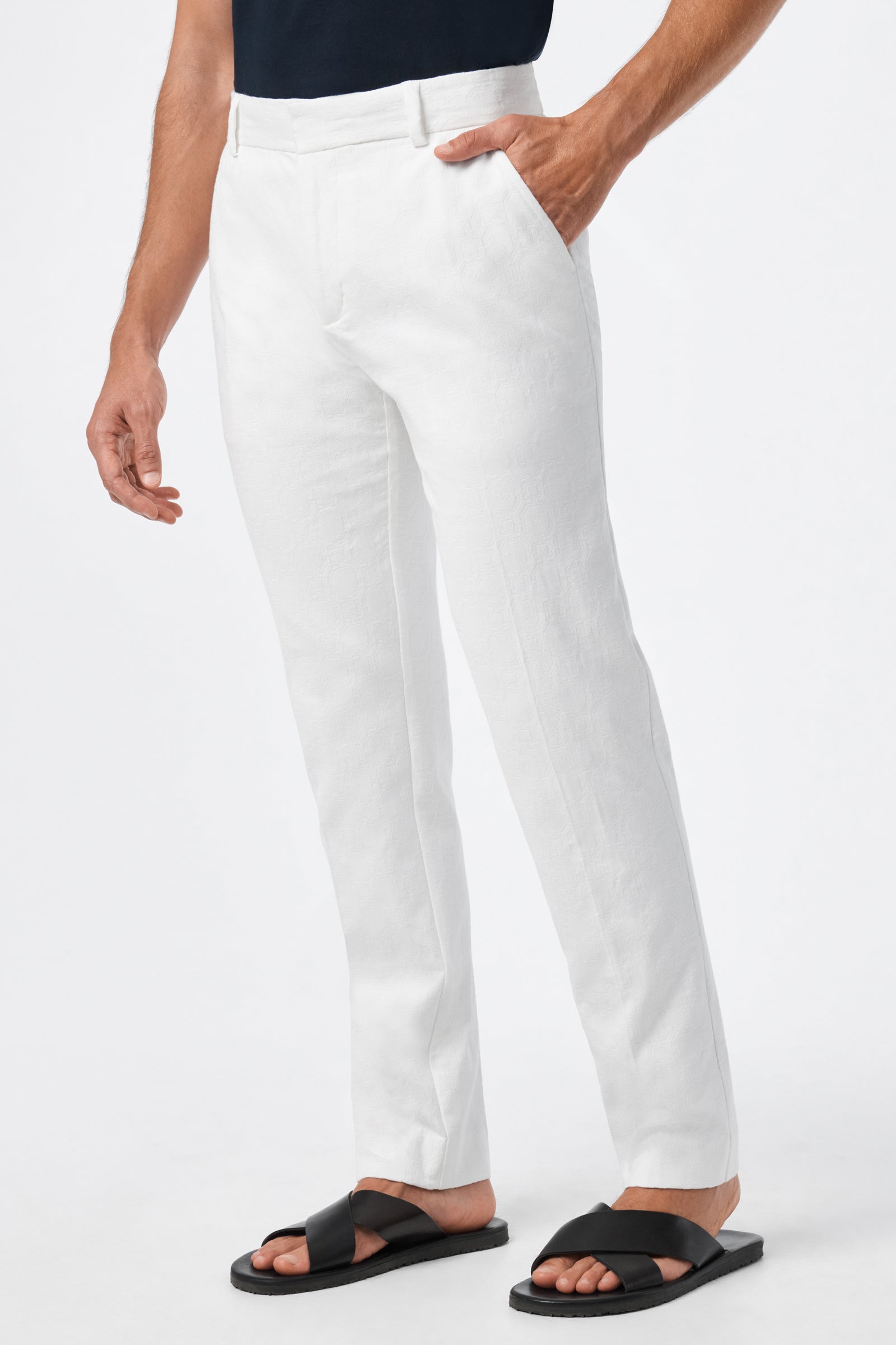 A person wearing Clyde Slim Trouser - Stretch Herringbone in white, paired with a tucked-in black shirt and black crisscross sandals, stands with one hand in their pocket against a plain white background.