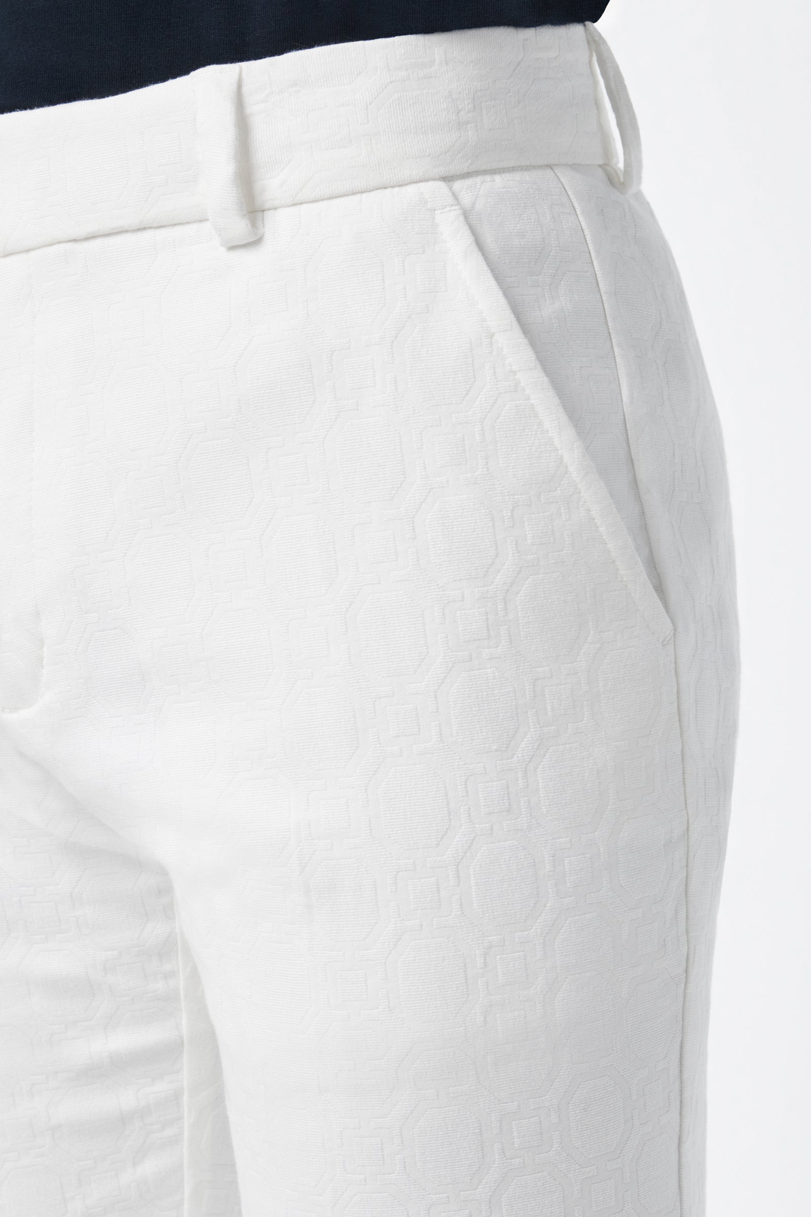 Close-up of a person wearing Clyde Slim Trouser - Stretch Herringbone in textured white with a subtle geometric pattern, side pocket, and belt-looped waistband; only the lower torso is shown against a plain background.