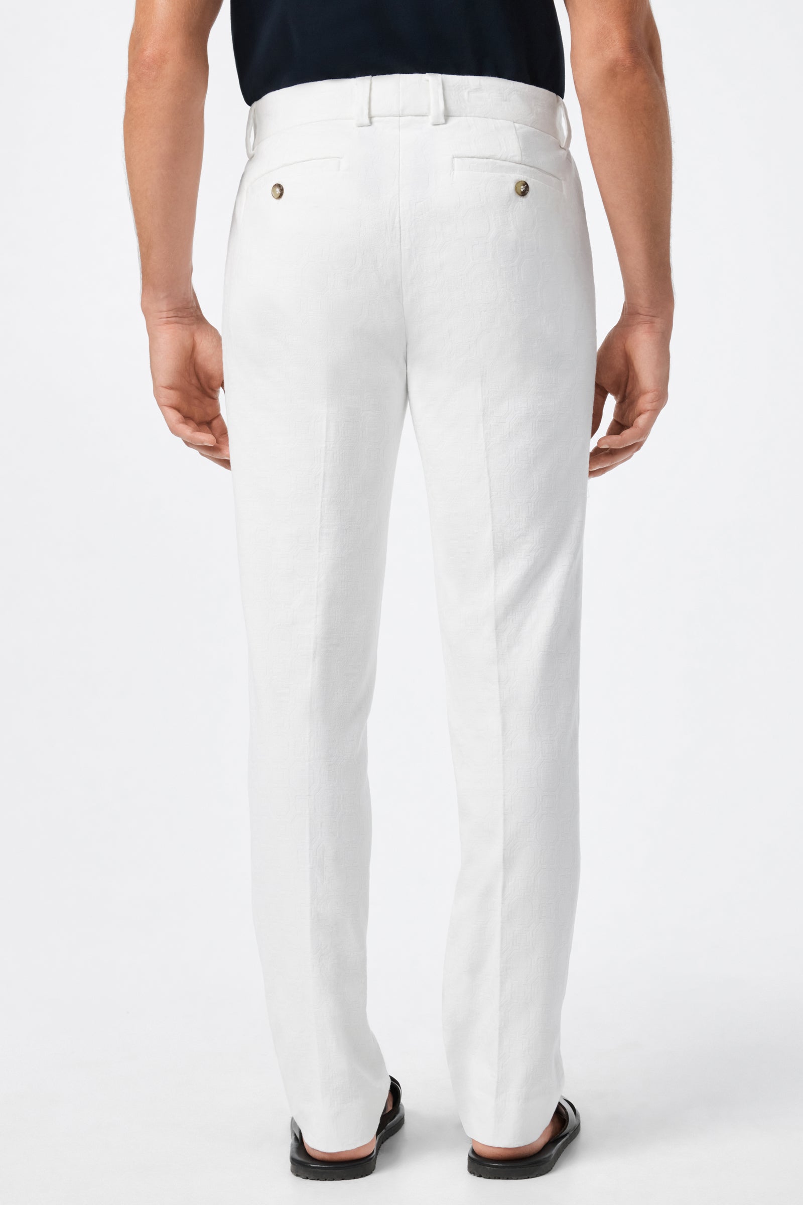 A man is shown from the back, wearing Clyde Slim Trouser - Stretch Herringbone in white, a tucked-in black shirt, and black shoes, standing against a plain white background.