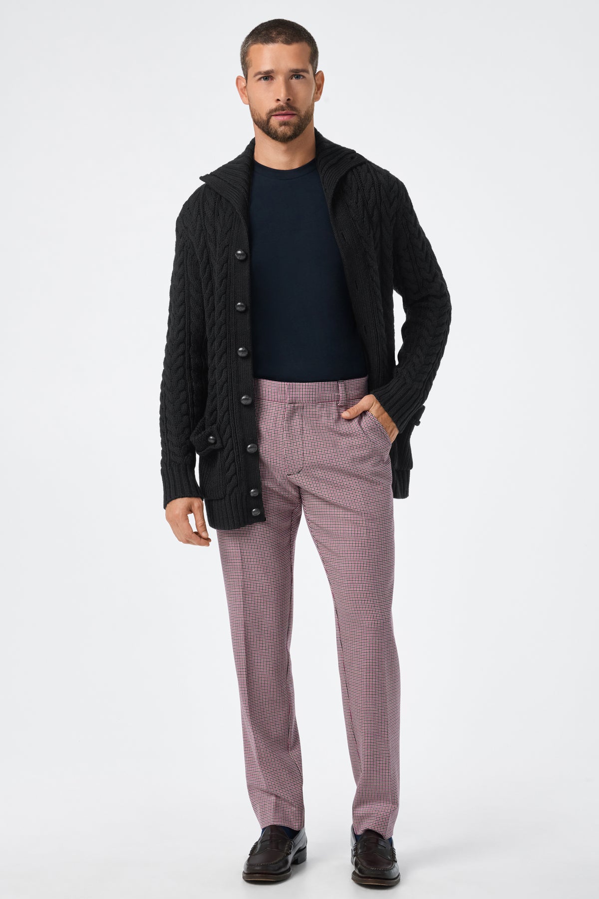 A man stands facing forward in the Raleigh Sweater Coat, a black cable-knit cardigan, styled with a navy shirt, pink checkered trousers, and dark brown dress shoes against a plain white background.
