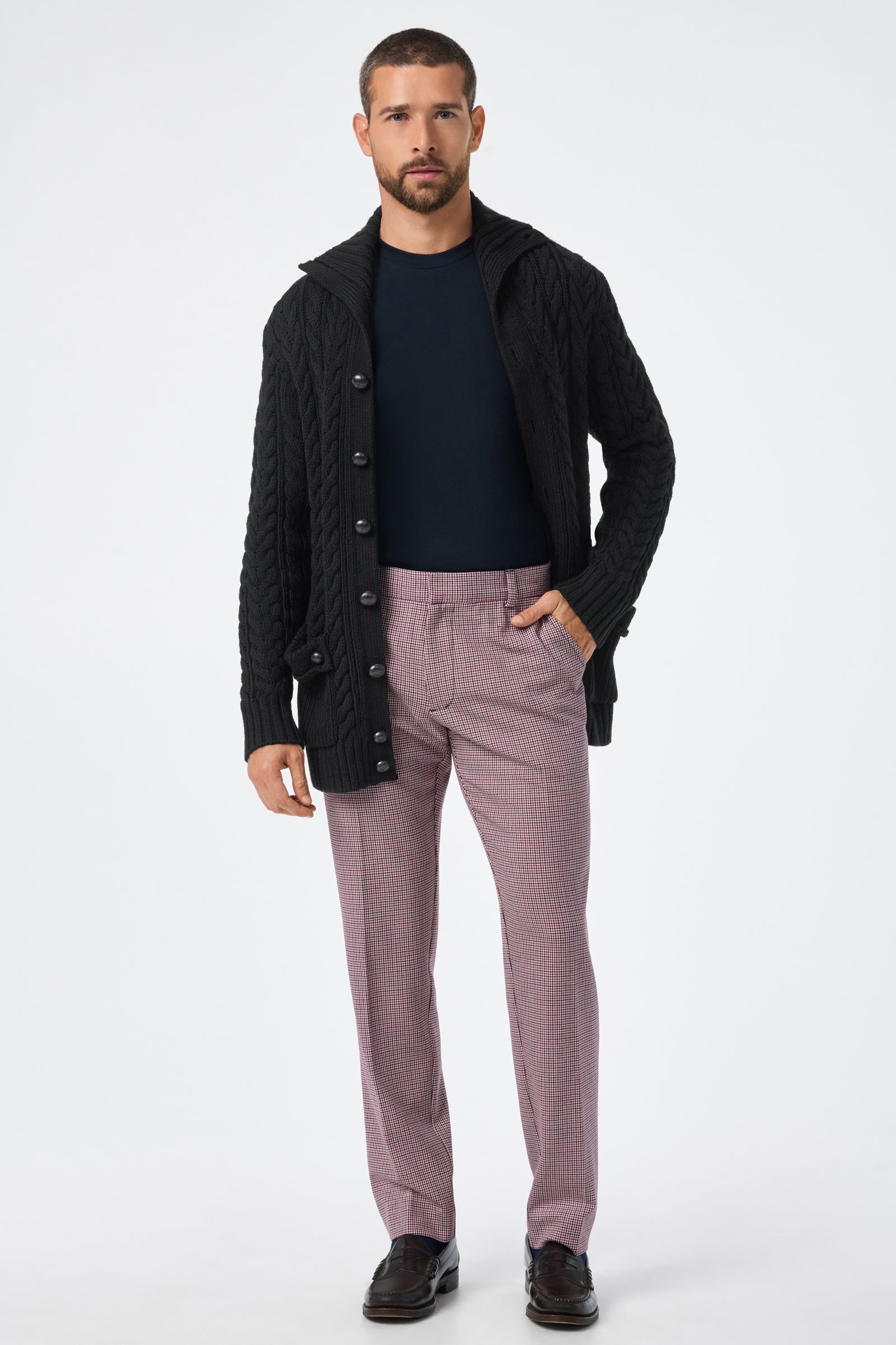 A man stands facing forward in the Raleigh Sweater Coat, a black cable-knit cardigan, styled with a navy shirt, pink checkered trousers, and dark brown dress shoes against a plain white background.