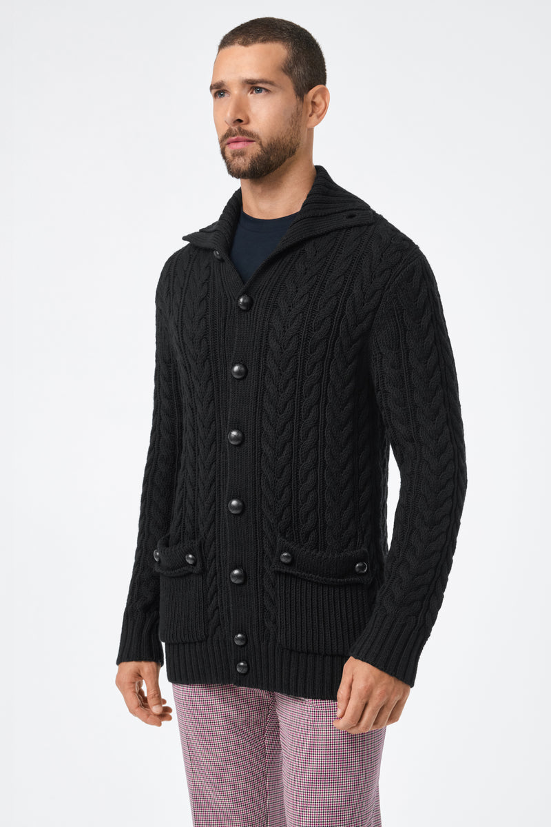 A man with short hair and a beard wears the Raleigh Sweater Coat—a black, chunky cable-knit button-up cardigan with front pockets—over a dark shirt and light plaid pants, standing against a plain white background.