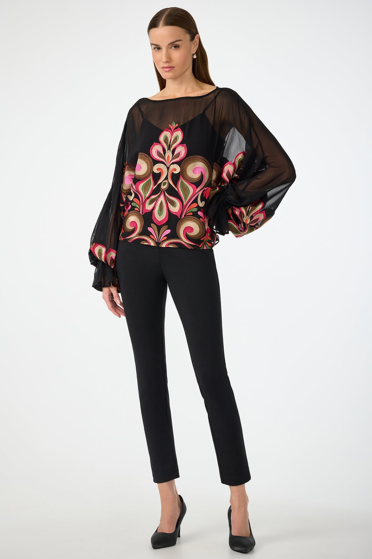 A woman poses against a white background wearing the Soho Embroidered Top by Sanctuary—a sheer black blouse with bold, colorful floral embroidery and voluminous sleeves—paired with fitted black pants and black heels.