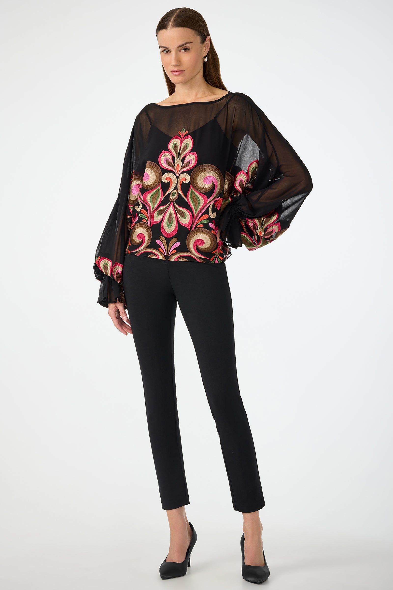 A woman poses against a white background wearing the Soho Embroidered Top by Sanctuary—a sheer black blouse with bold, colorful floral embroidery and voluminous sleeves—paired with fitted black pants and black heels.