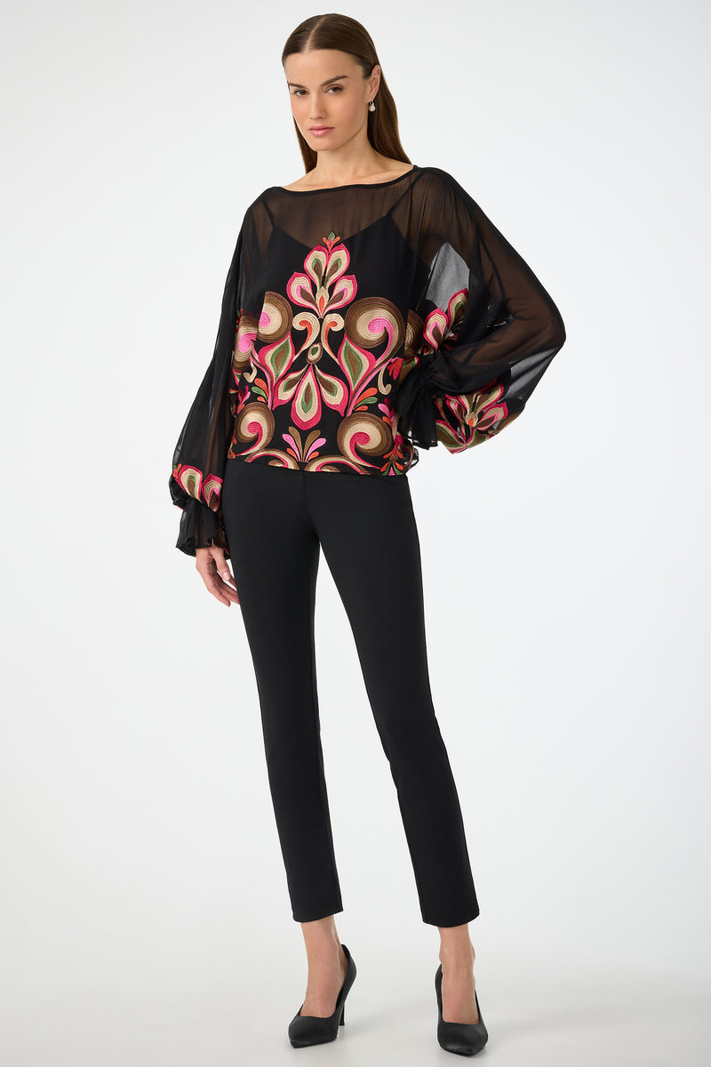 A woman poses against a white background wearing the Soho Embroidered Top by Sanctuary—a sheer black blouse with bold, colorful floral embroidery and voluminous sleeves—paired with fitted black pants and black heels.
