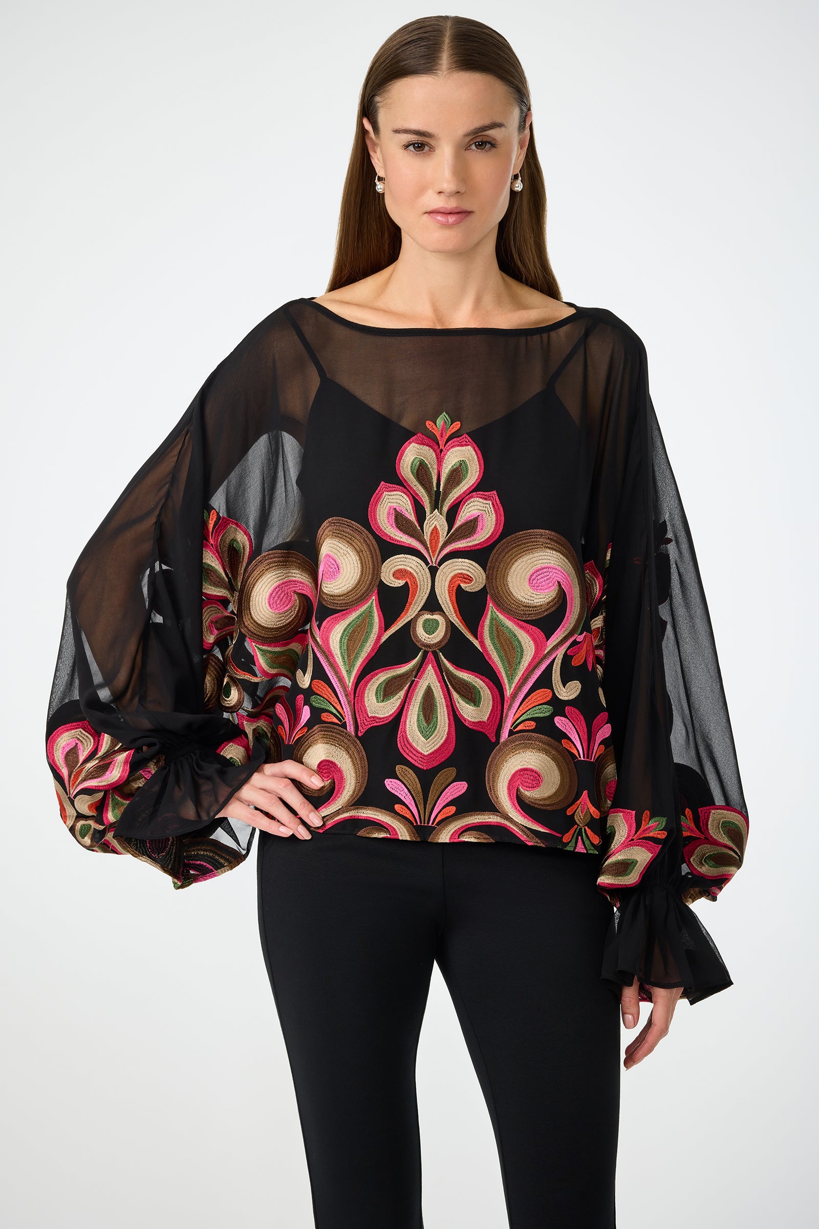 A woman wears the Sanctuary Soho Embroidered Top, a sheer blouse with vibrant pink, green, and gold embroidery and voluminous sleeves, styled over black pants against a plain light background.