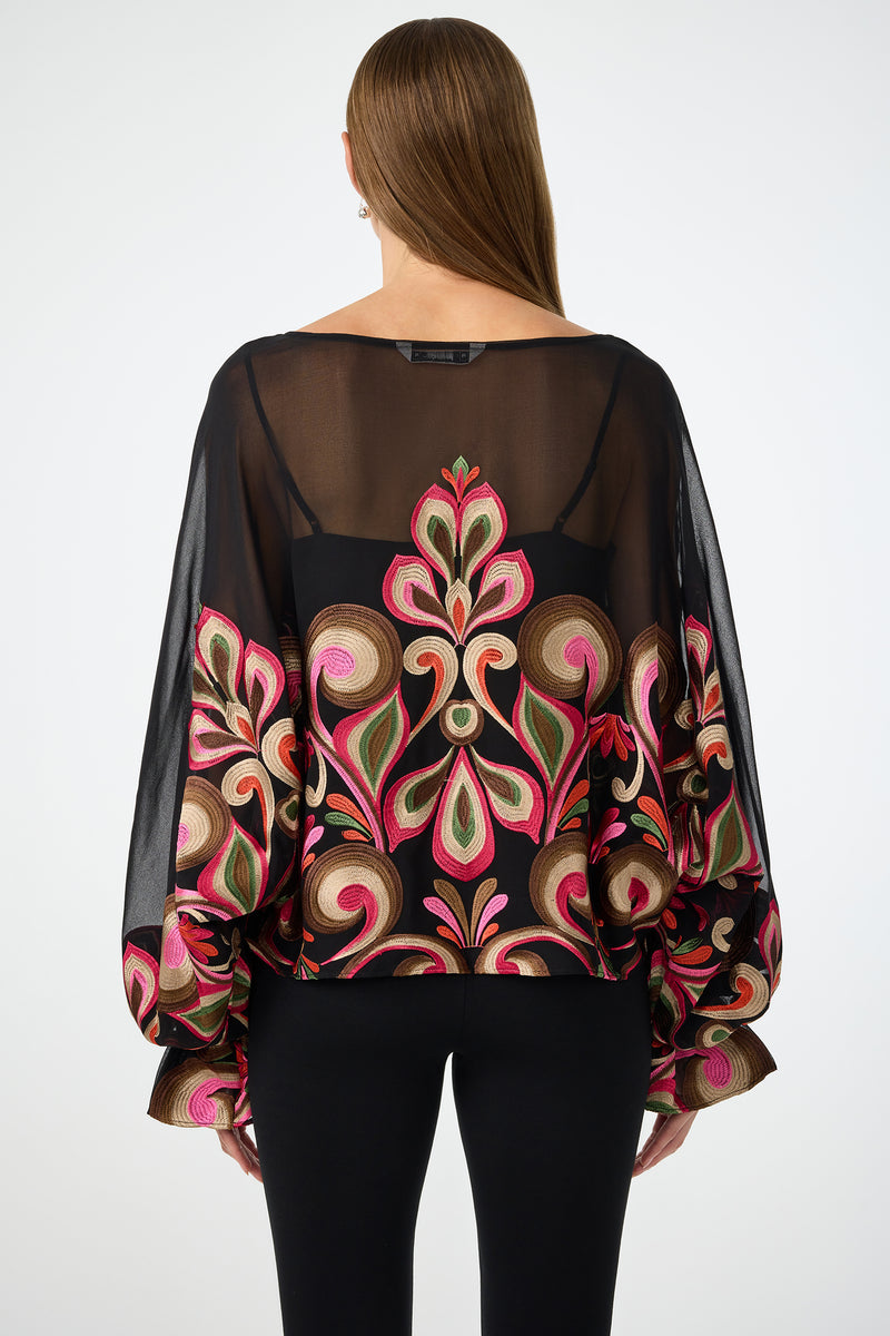 A woman with straight brown hair faces away from the camera, showcasing the back of the Sanctuary Soho Embroidered Top—a sheer black blouse with colorful floral embroidery and billowy sleeves—paired with black pants.