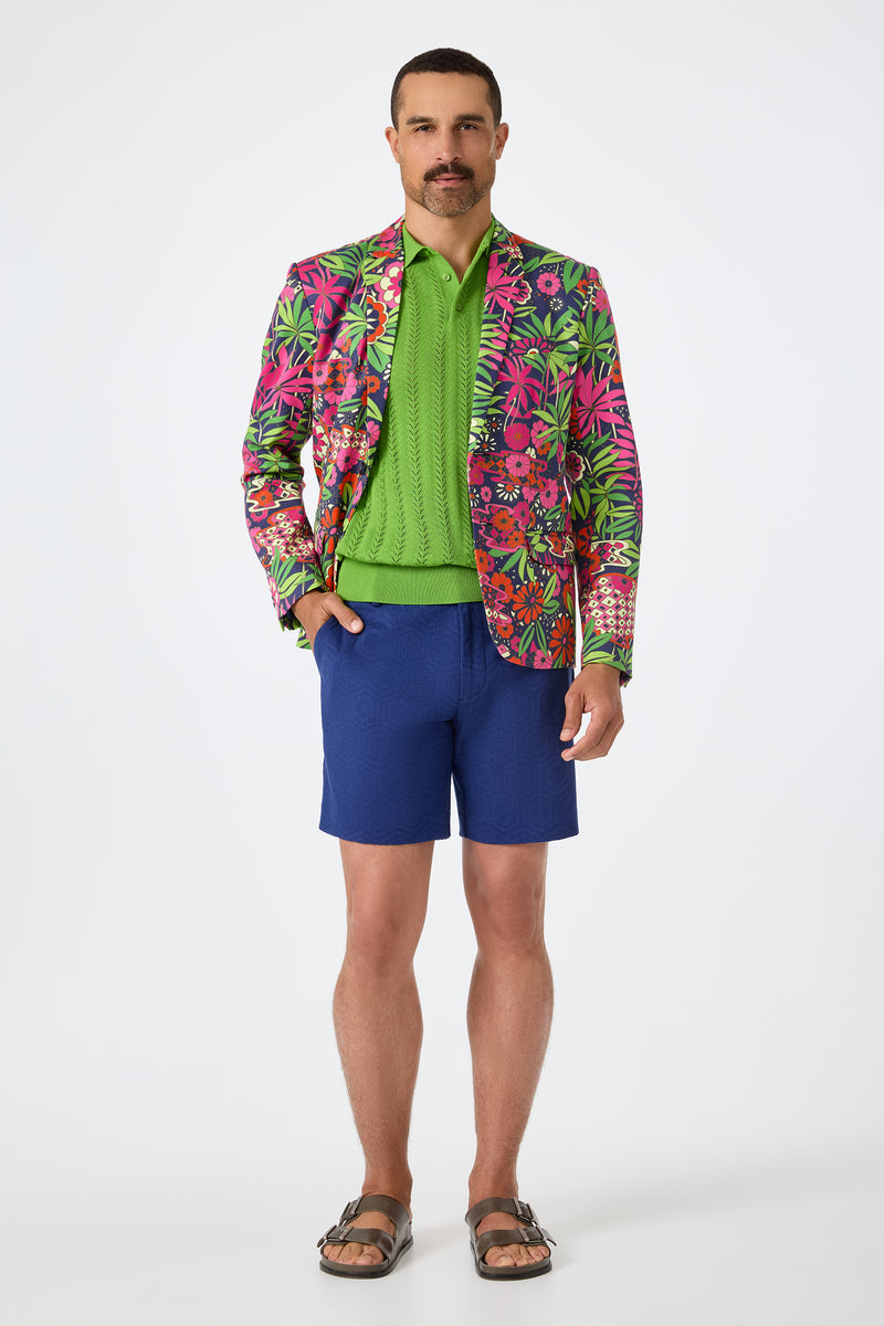 A man with a mustache stands against a white background wearing the Thurston Blazer - Fumi Floral, a bright green sweater, blue shorts, and brown sandals.