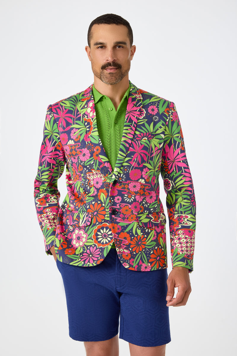A man stands against a plain white background wearing the Thurston Blazer in Fumi Floral over a green polo shirt and blue shorts.