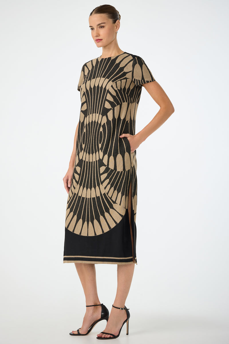 A woman wearing the Sugarita Midi Dress - Juniper Fil Coupe, featuring short sleeves, knee-length fit, bold black and beige geometric pattern with side slit and black hem, paired with black heels against a plain light background.