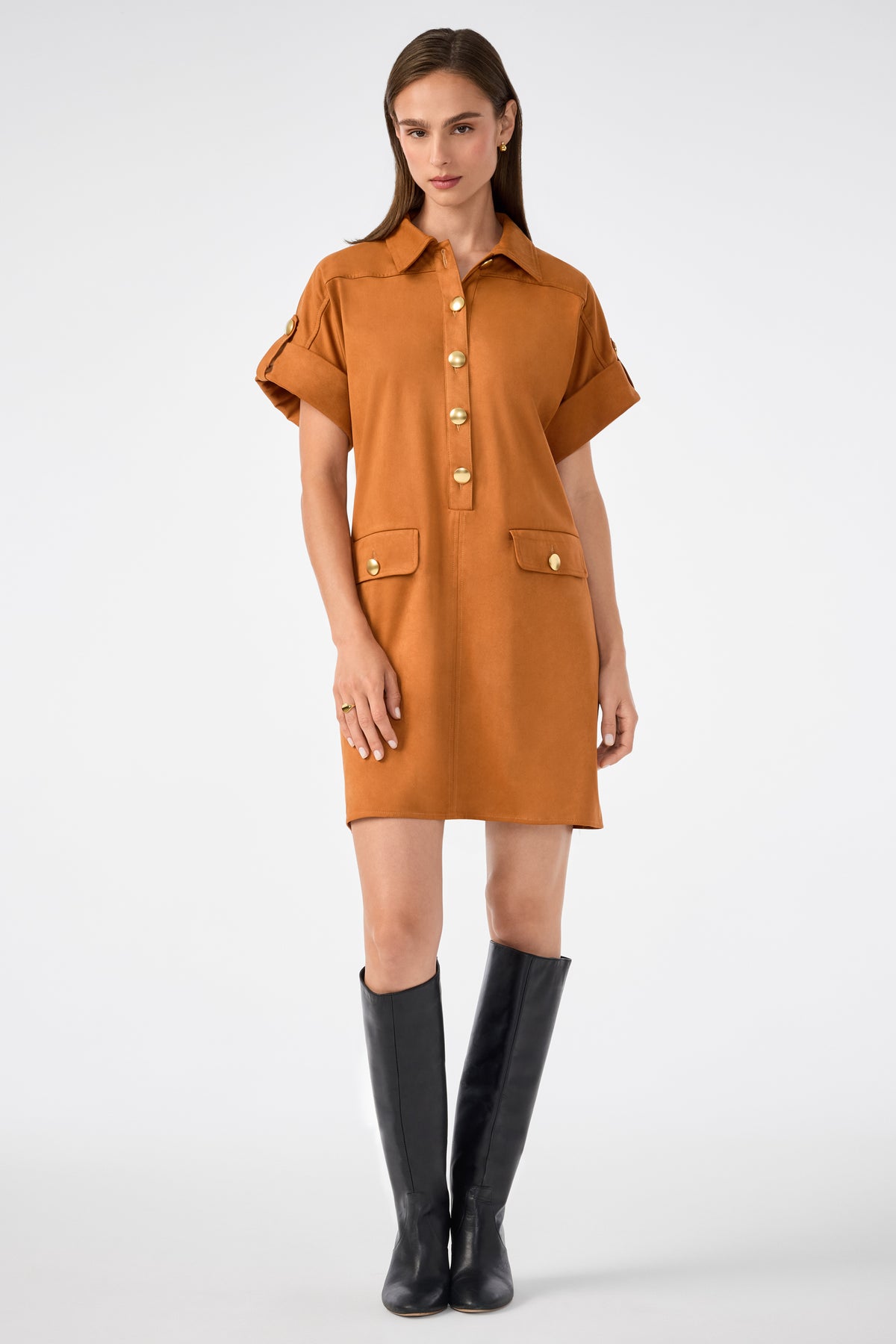 A woman wears the Sequoia Dress, a short-sleeved, button-front brown shirt dress with large gold buttons and front pockets, styled with knee-high black boots against a plain white background.