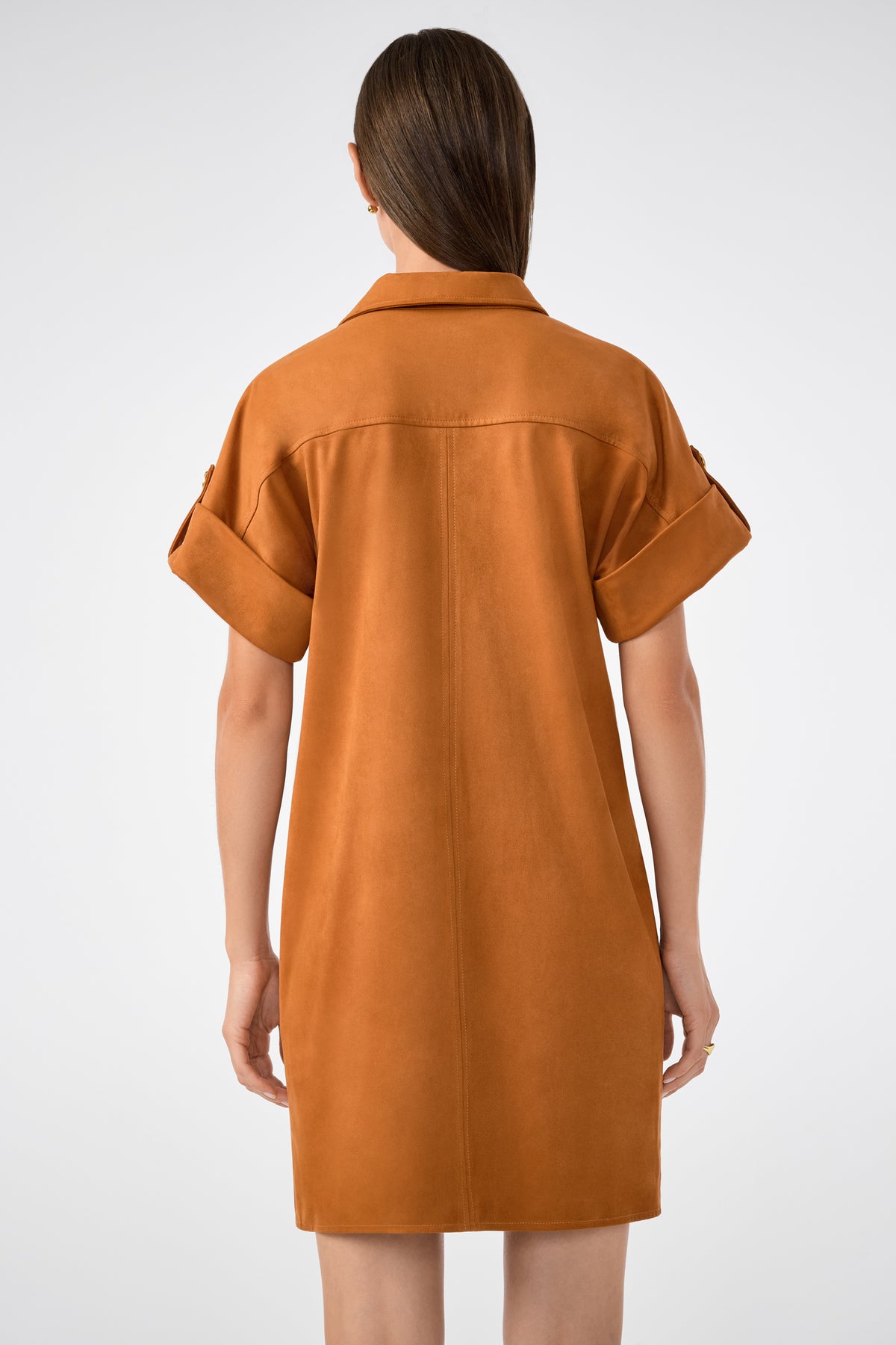 A person with long brown hair is seen from the back wearing the Sequoia Dress—a loose, short-sleeved caramel brown dress with rolled cuffs—against a plain white background.