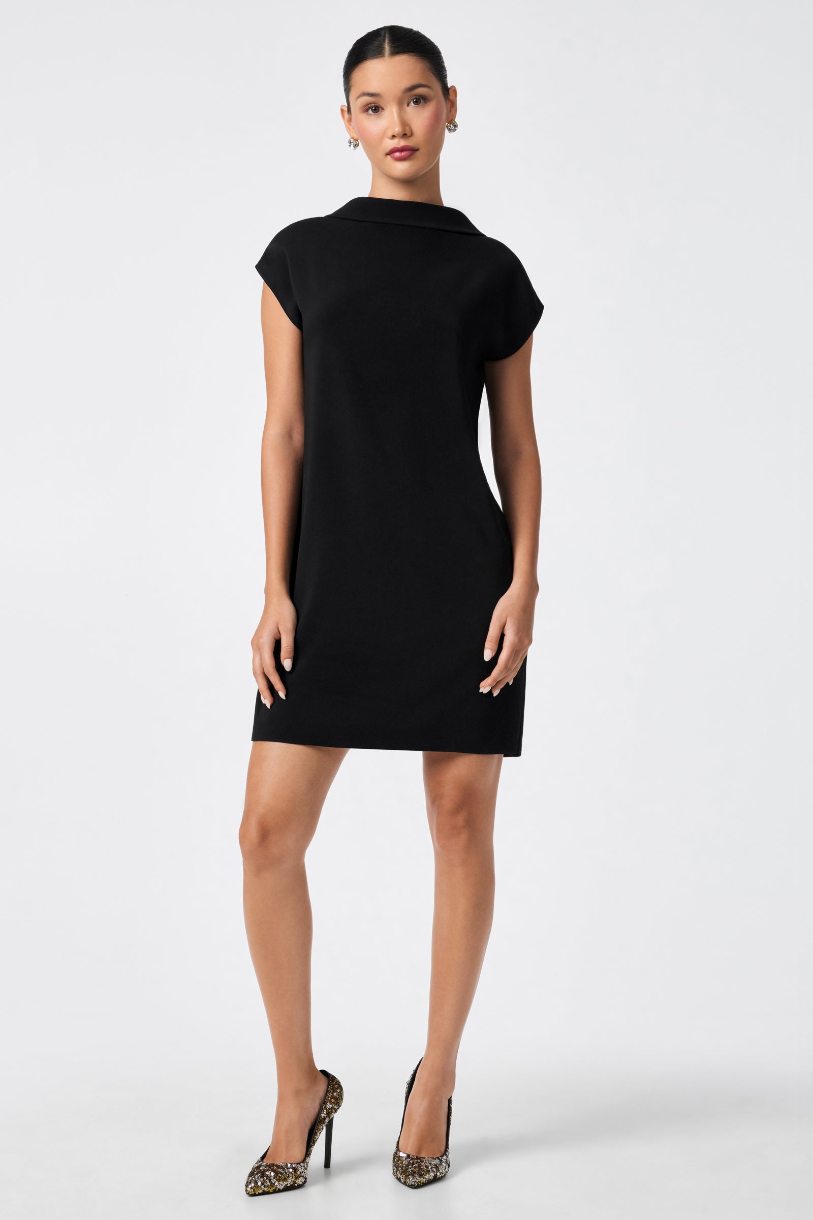 A woman stands against a plain light background wearing the CHORUS 2 DRESS, a short-sleeved, knee-length black dress, paired with leopard print high heels. She looks forward with her arms relaxed at her sides.