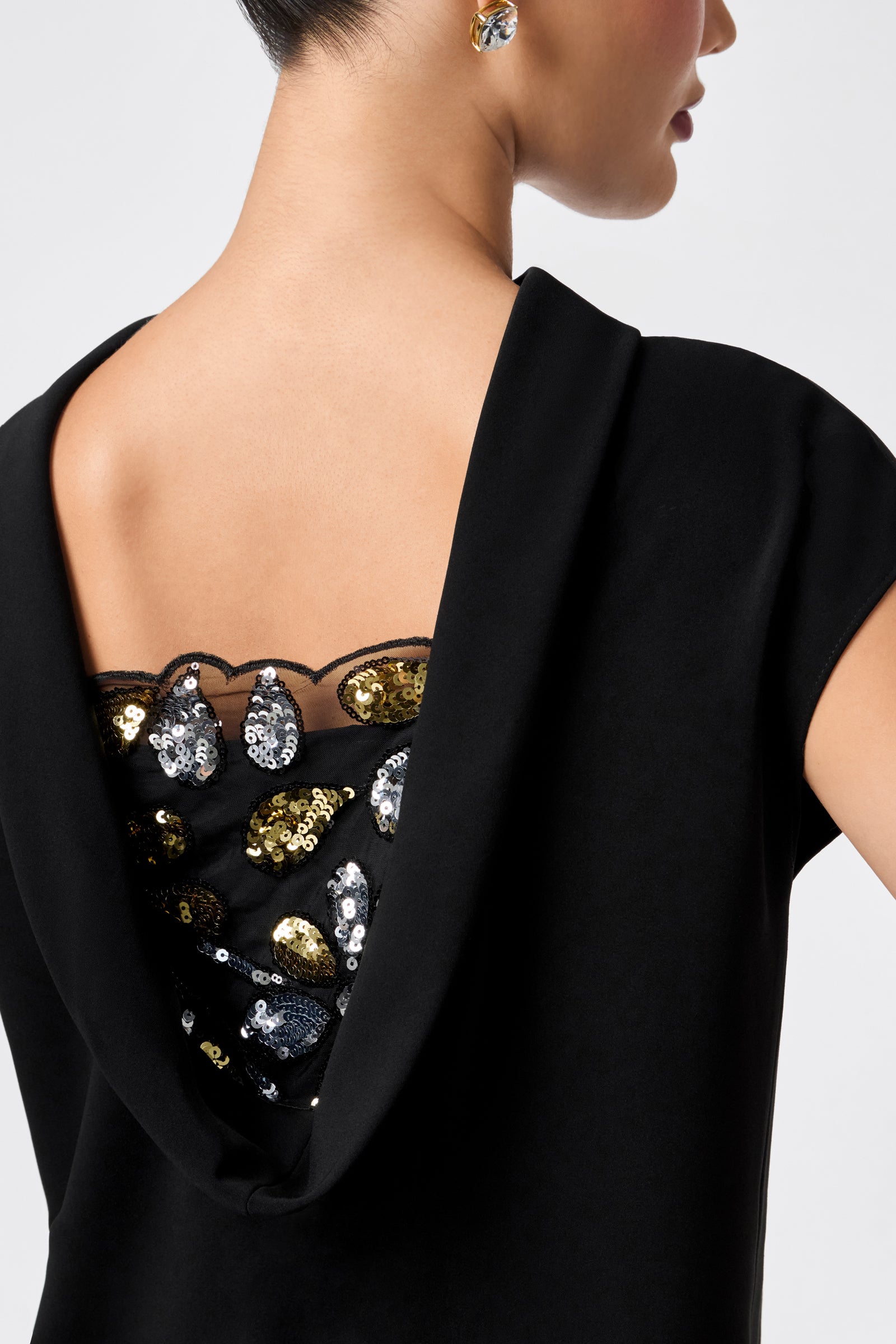 A woman with short hair and a small earring wears the CHORUS 2 DRESS, which features a black top with a deep cowl back, revealing sheer fabric adorned with gold, silver, and black sequin embroidery.
