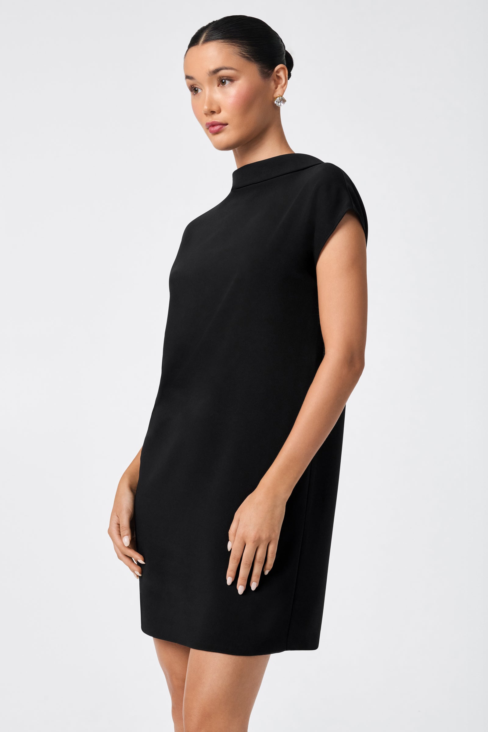 A woman poses in the CHORUS 2 DRESS, a simple and elegant black short-sleeve dress with a high neckline, against a plain light gray background.