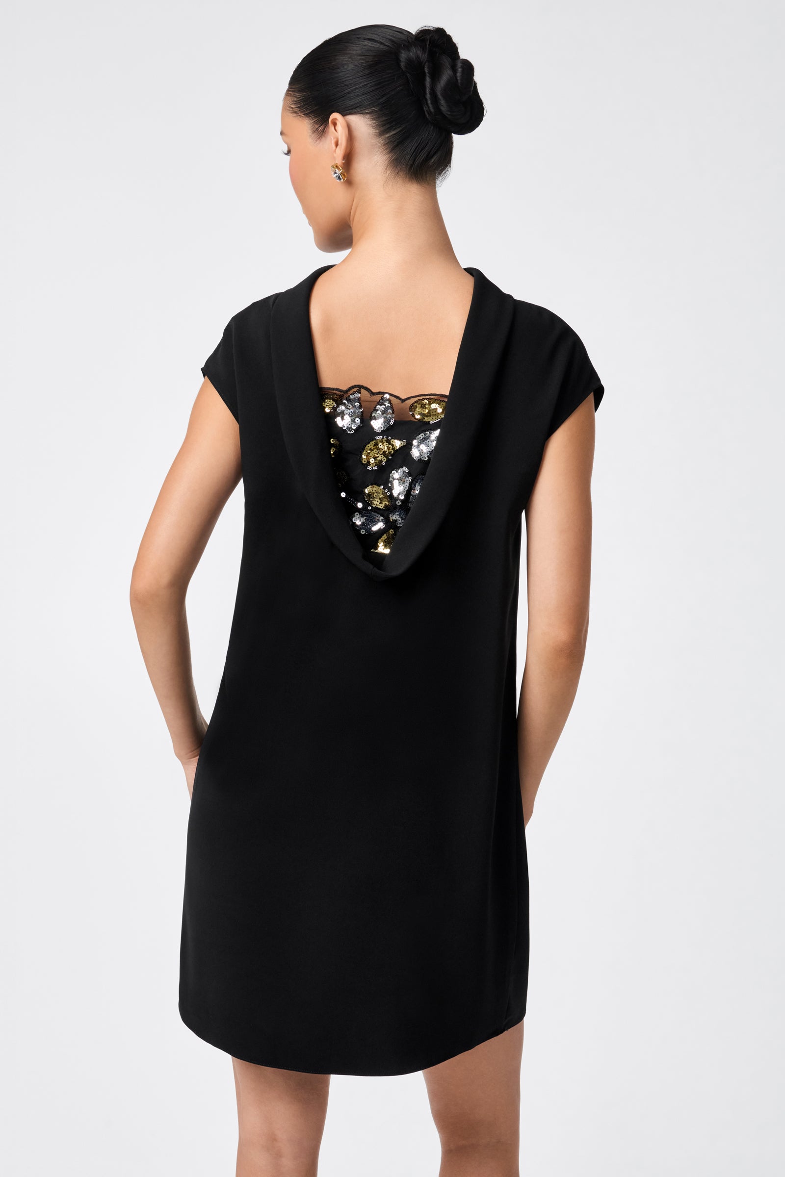 A woman stands with her back to the camera, wearing the CHORUS 2 DRESS—a black gown featuring a draped cowl back embellished with shiny gold and silver sequins. Her dark hair is styled in a neat updo.