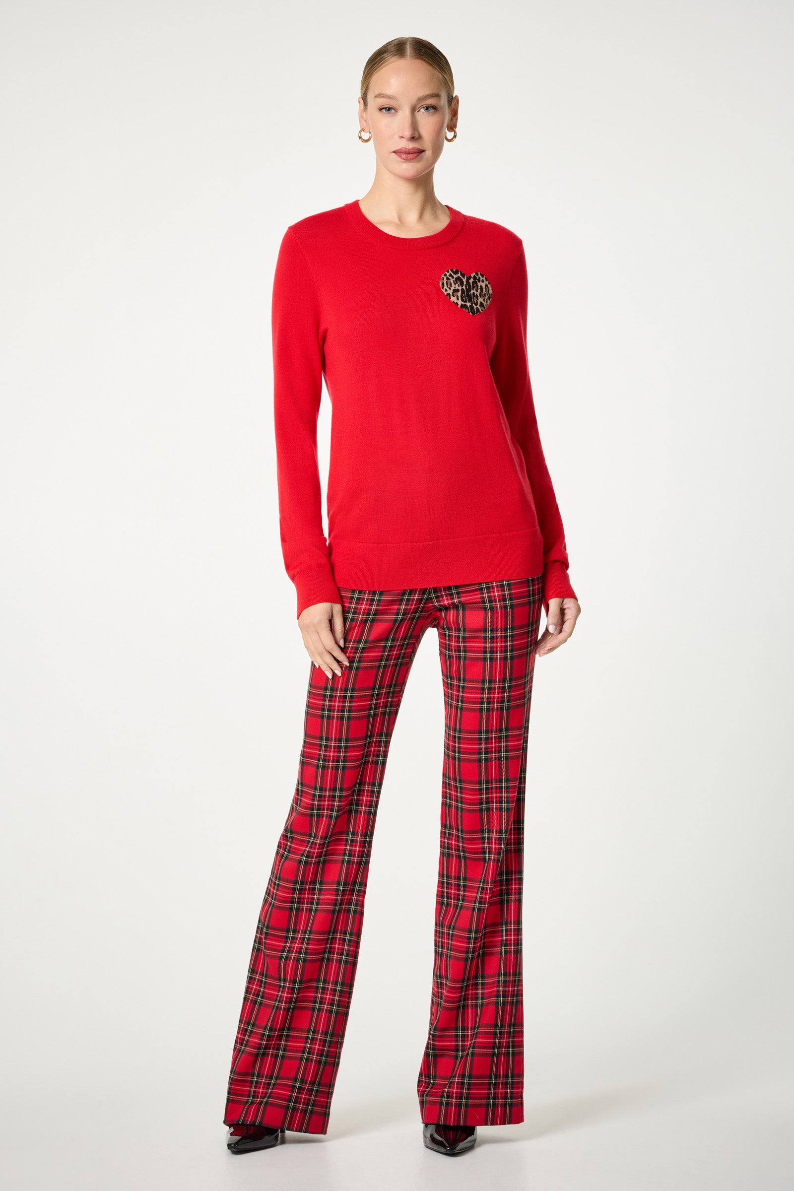 A woman stands against a white background wearing the Jaguar Heart Crewneck Sweater, featuring a leopard-print heart on the chest, paired with red plaid flared pants, black heels, and gold hoop earrings.
