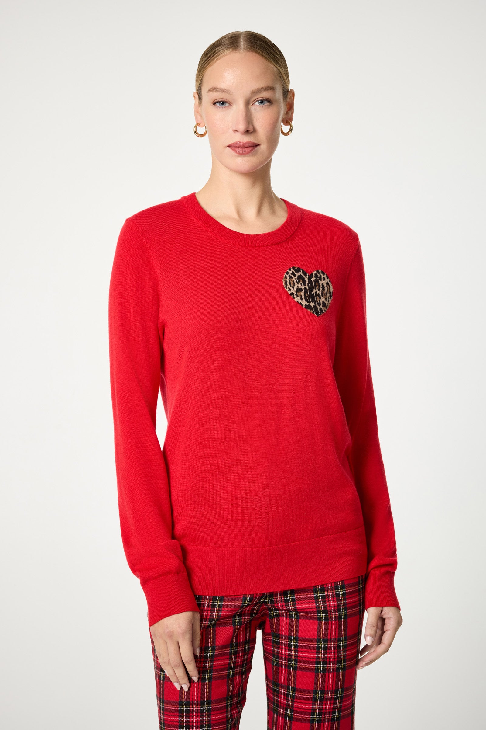 A woman wears the Jaguar Heart Crewneck Sweater—bright red with a leopard print heart patch on the left chest—paired with red plaid pants and gold hoop earrings. Her hair is pulled back and the background is plain white.
