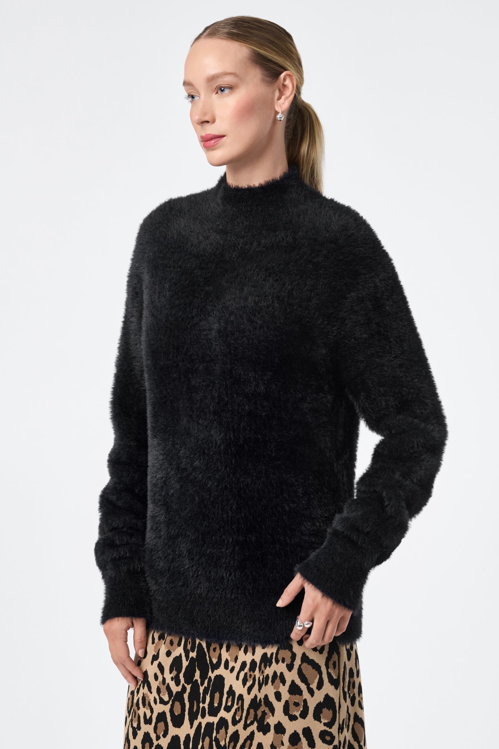 A woman with light skin and long, straight hair tied back wears the Bazaar Pullover, a fuzzy black sweater, paired with a leopard print skirt. She stands against a plain light background, gazing slightly to the side with a neutral expression.