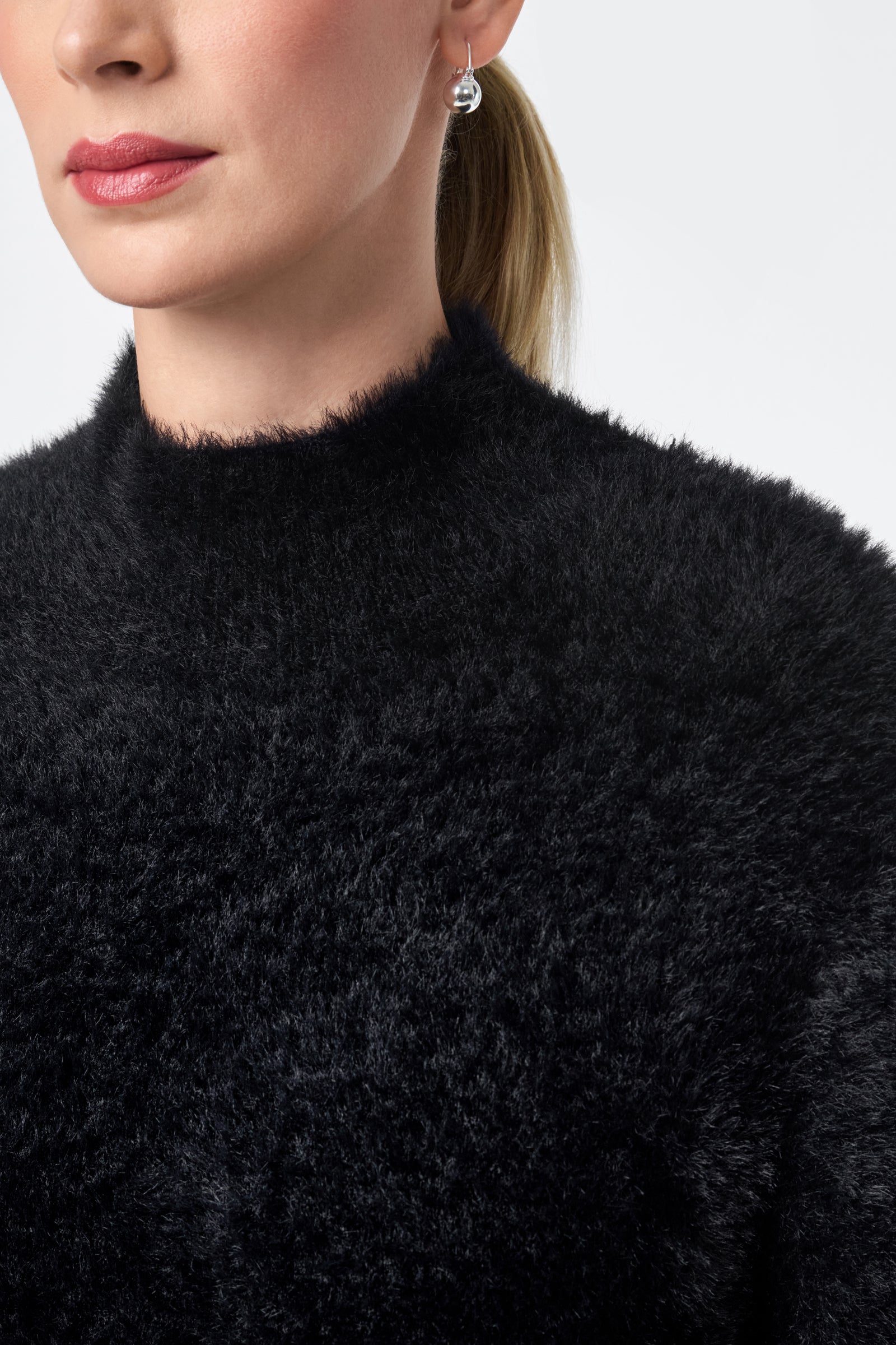 A woman with light skin wears the Bazaar Pullover, a black fuzzy sweater. Her hair is tied back in a ponytail, and she has a single silver drop earring. Only the lower half of her face and upper torso are visible.