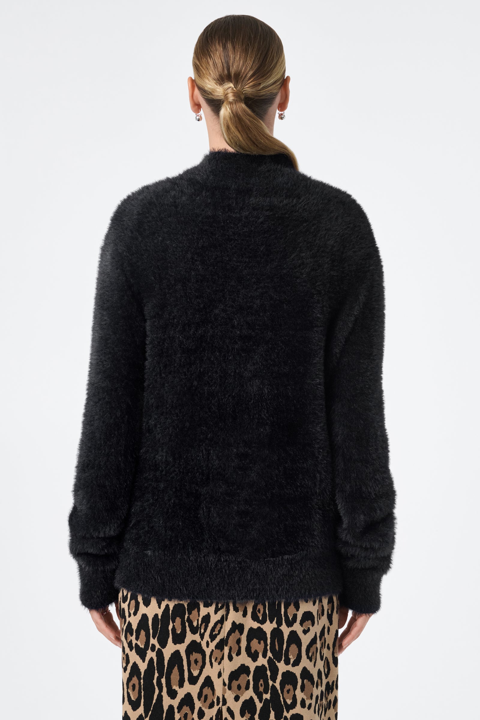 A person with blonde hair in a low ponytail is shown from the back wearing the black fuzzy Bazaar Pullover and a tan leopard print skirt, set against a plain white background.