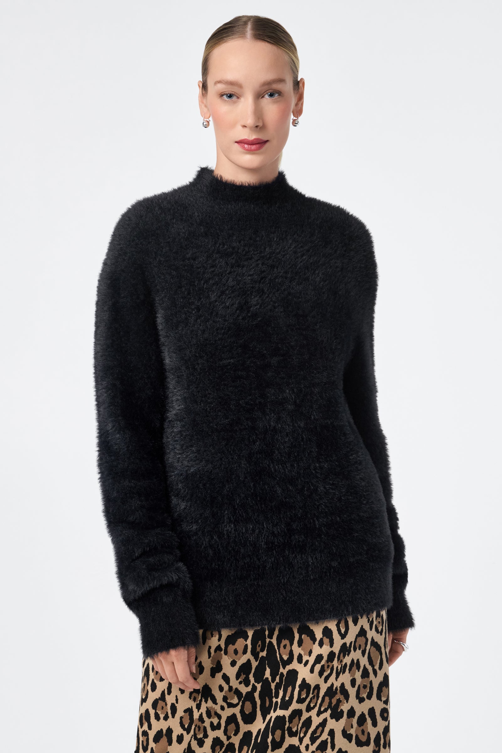 A woman with light skin and slicked-back hair models the Bazaar Pullover, a fuzzy black sweater, paired with a leopard print skirt against a plain, light background.