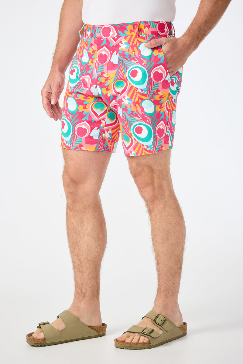 A man stands with his hands in his pockets against a plain white background, wearing a white shirt, beige sandals, and the Lawrence Short - Dramont Herringbone featuring pink, teal, and orange patterned shapes.