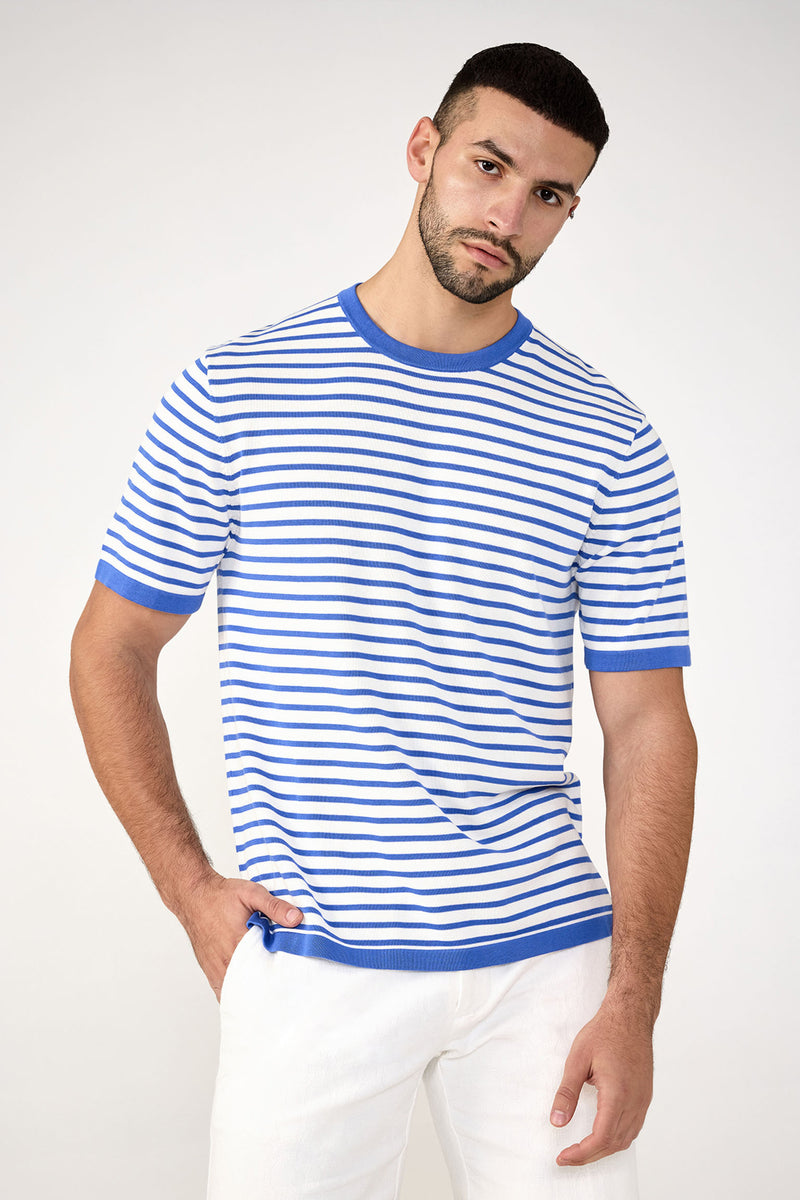 A man with short dark hair and a trimmed beard wears the Giovanni Stripe Short-Sleeve Pullover in Beach Ball Blue, paired with white pants. He stands with one hand in his pocket against a plain light background.