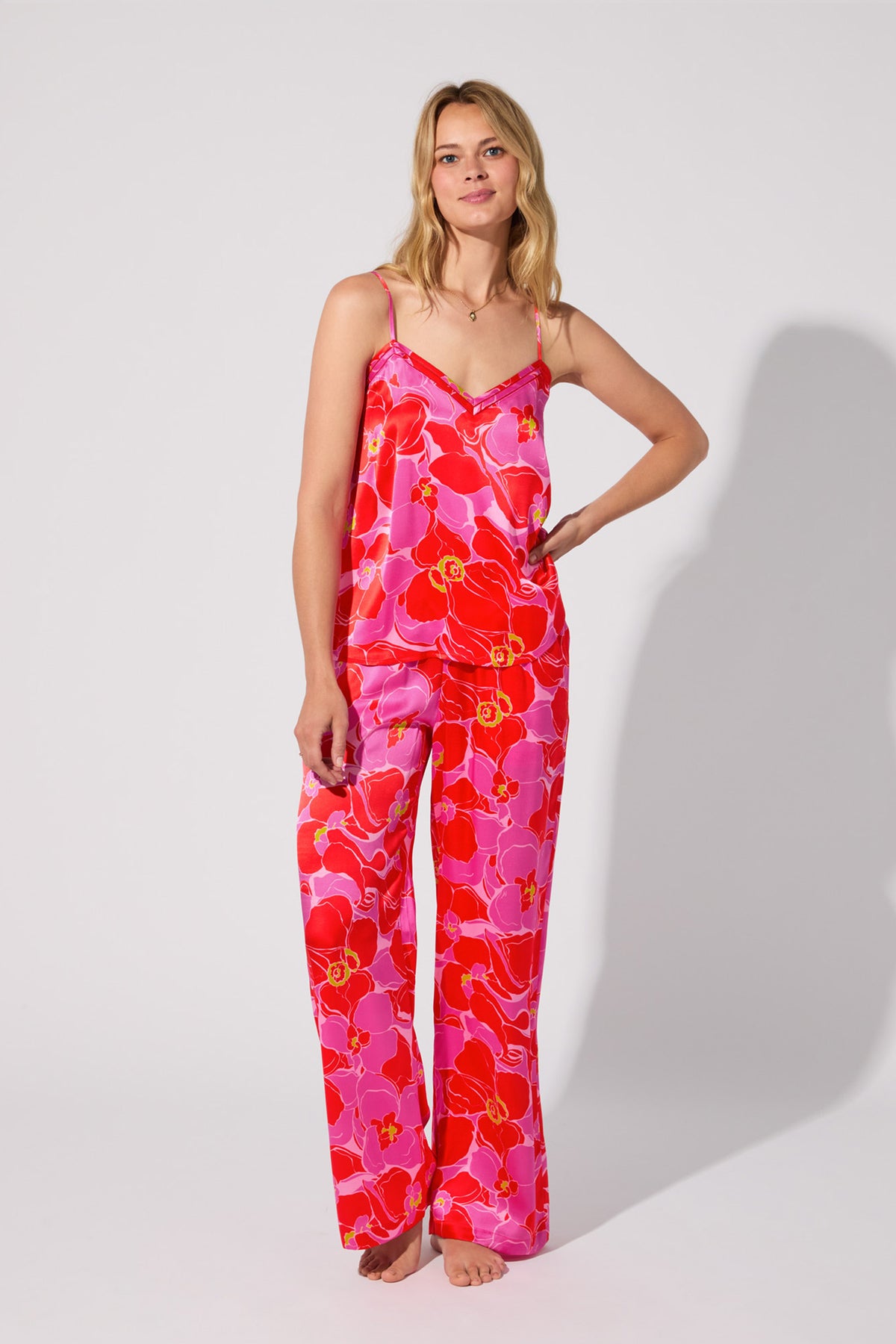 A woman poses barefoot against a plain white background, smiling slightly in the Silk Cami PJ Set – Winter Orchids, featuring a colorful floral satin pattern with a sleeveless camisole and loose pants.