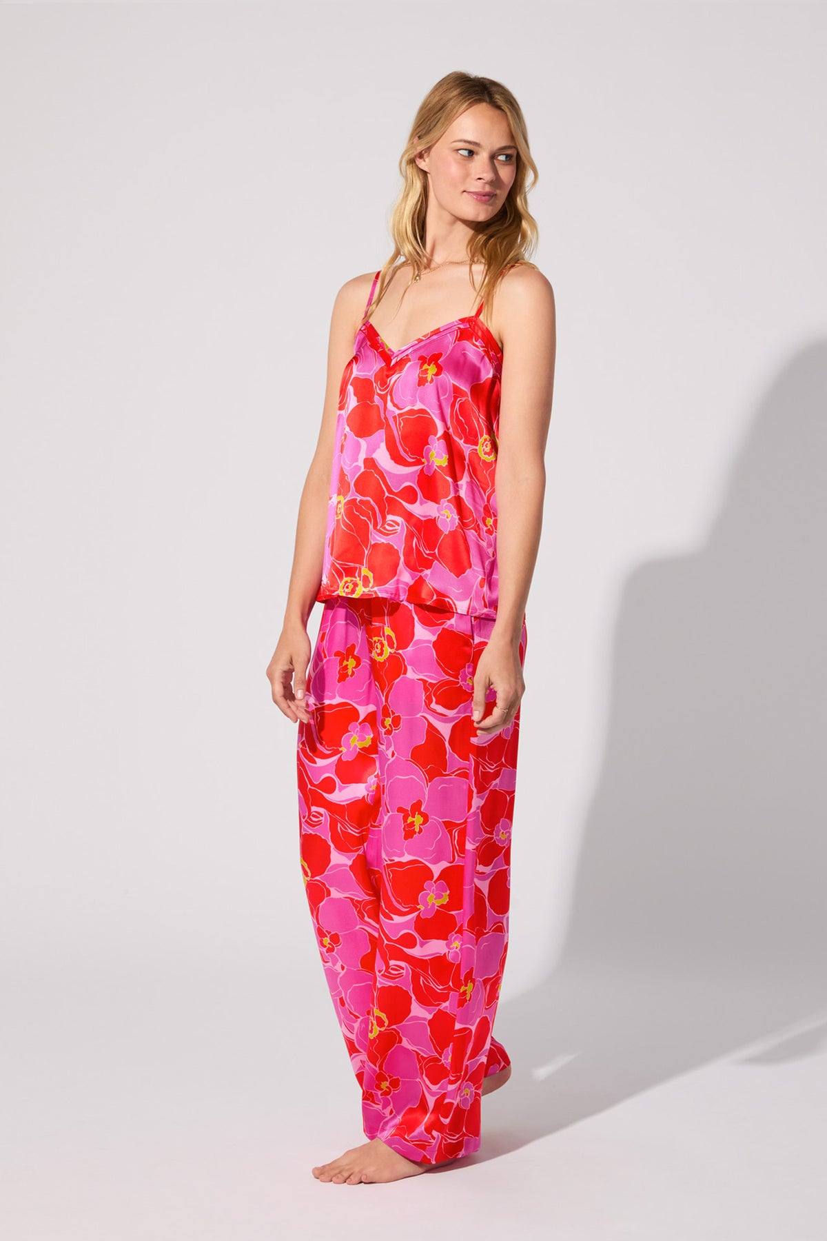 A woman stands barefoot in the Silk Cami PJ Set - Winter Orchids, featuring a matching pink and red floral sleeveless top and loose pants, set against a plain light background.