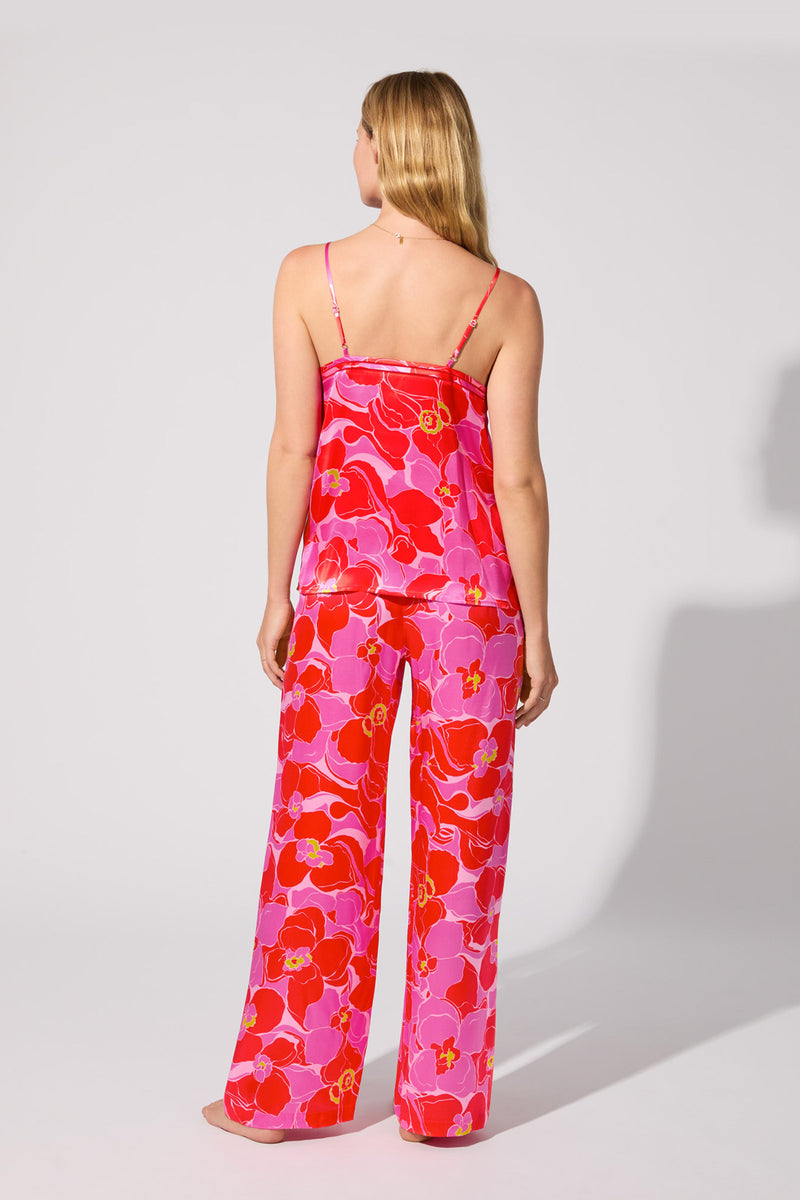 A woman with long blonde hair faces away, wearing the Silk Cami PJ Set - Winter Orchids, featuring a strappy top and loose pants with a bold pink, red, and purple floral print. The background is plain light gray.
