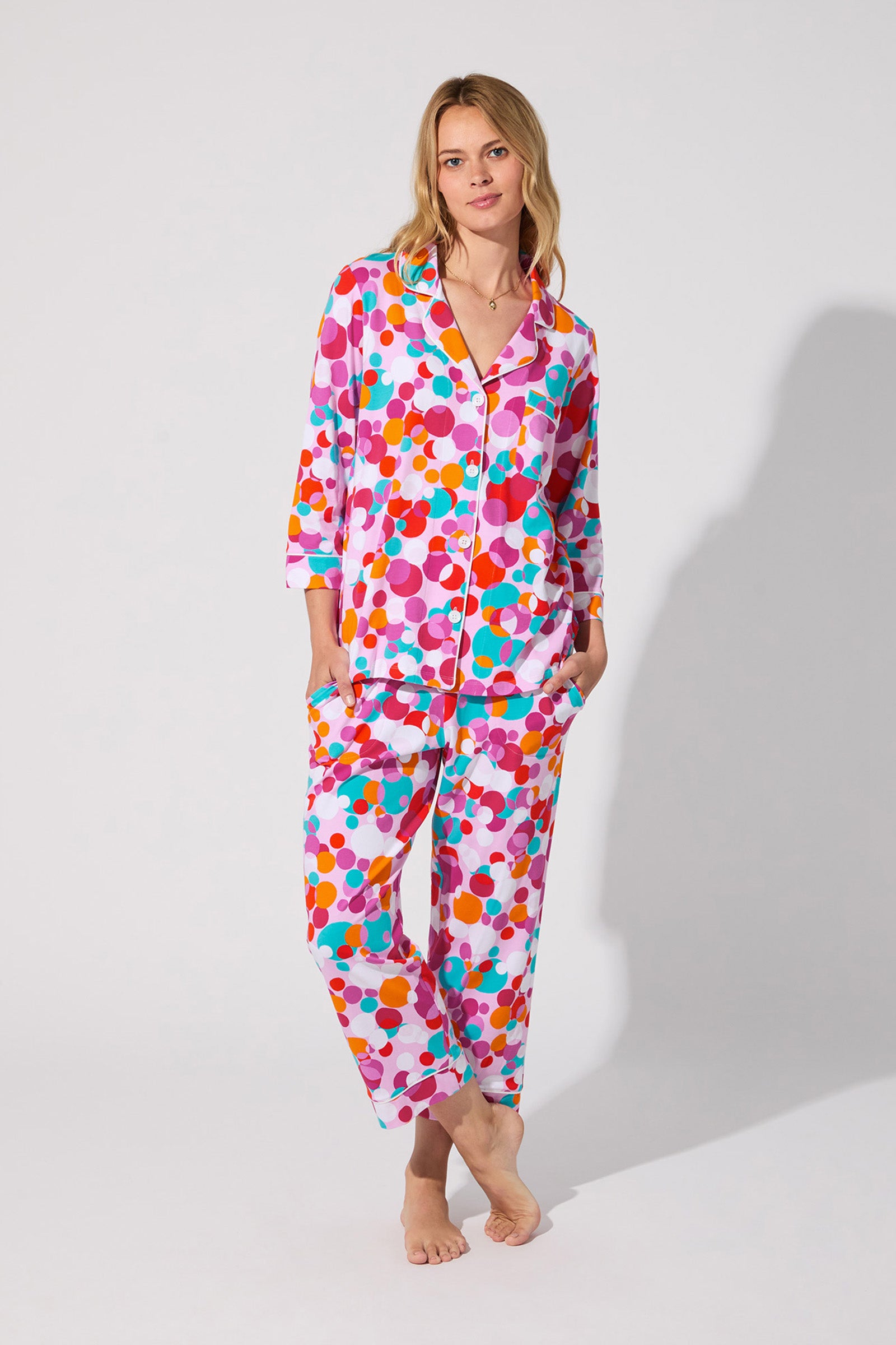 A woman stands barefoot in the Bubble Dots Crop Pj Set—colorful polka-dot patterned, button-up top and matching pants—against a plain light background.