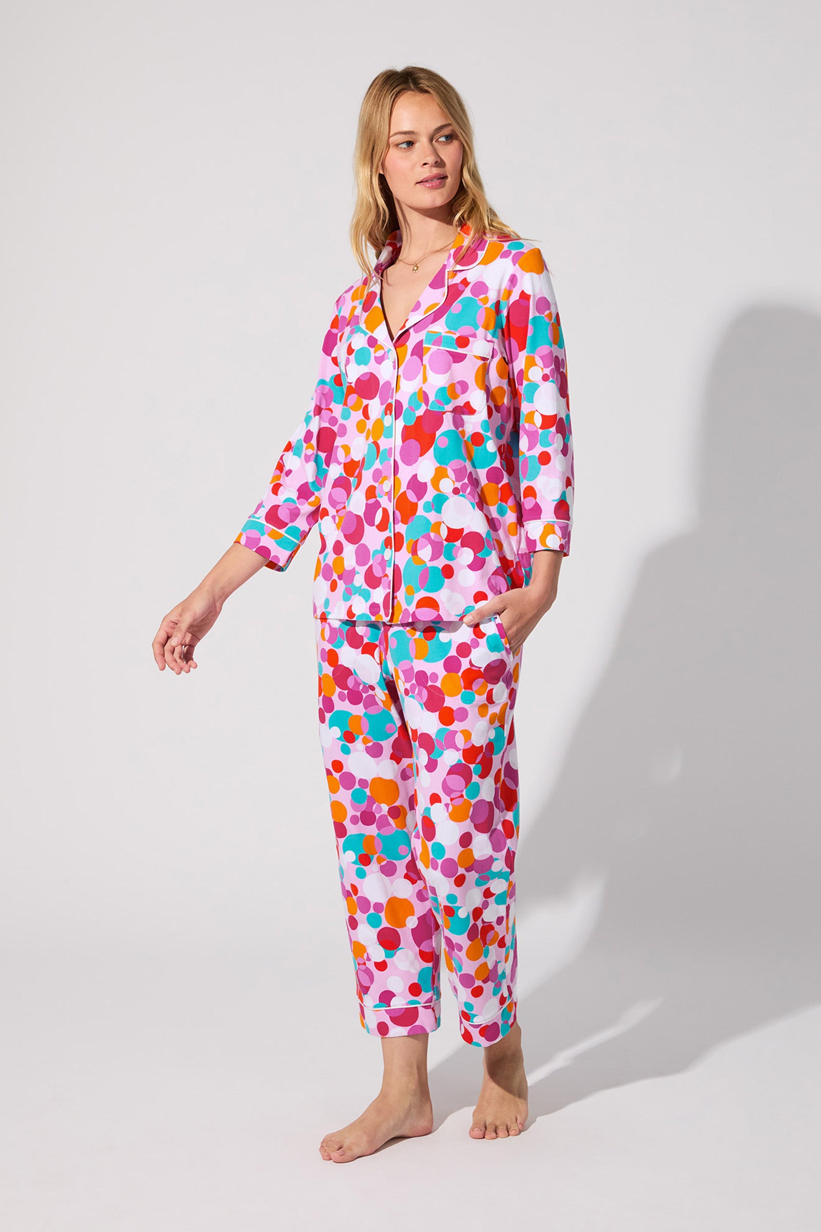 A woman stands barefoot in the Bubble Dots Crop Pj Set, featuring colorful polka dots, a button-up crop top, and long pants. She poses against a plain light background, looking slightly to the side with a relaxed expression.