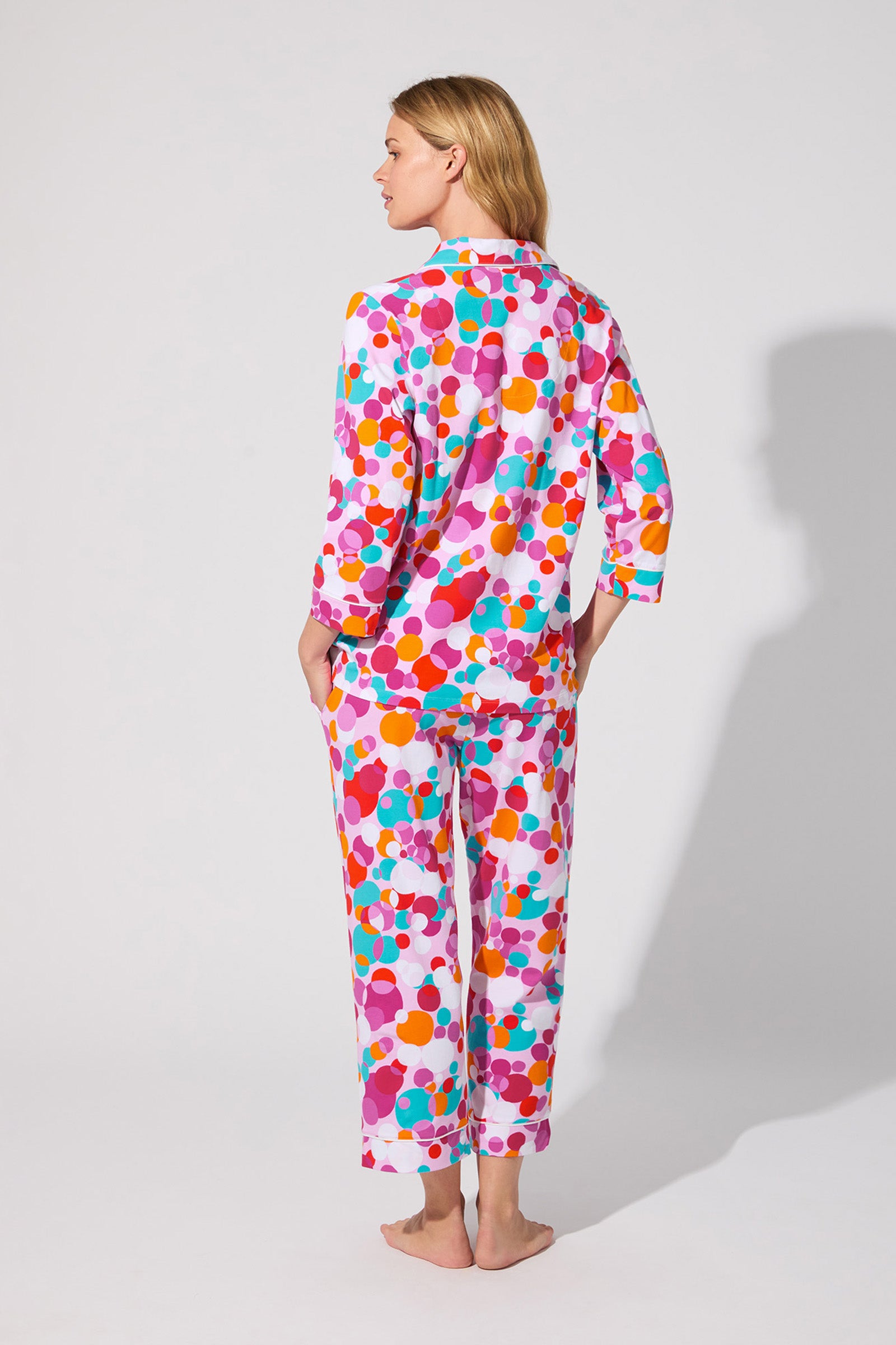 A person stands barefoot facing away, wearing the Bubble Dots Crop Pj Set featuring a bright, multicolored polka dot pattern in pink, orange, red, and purple on a white background.