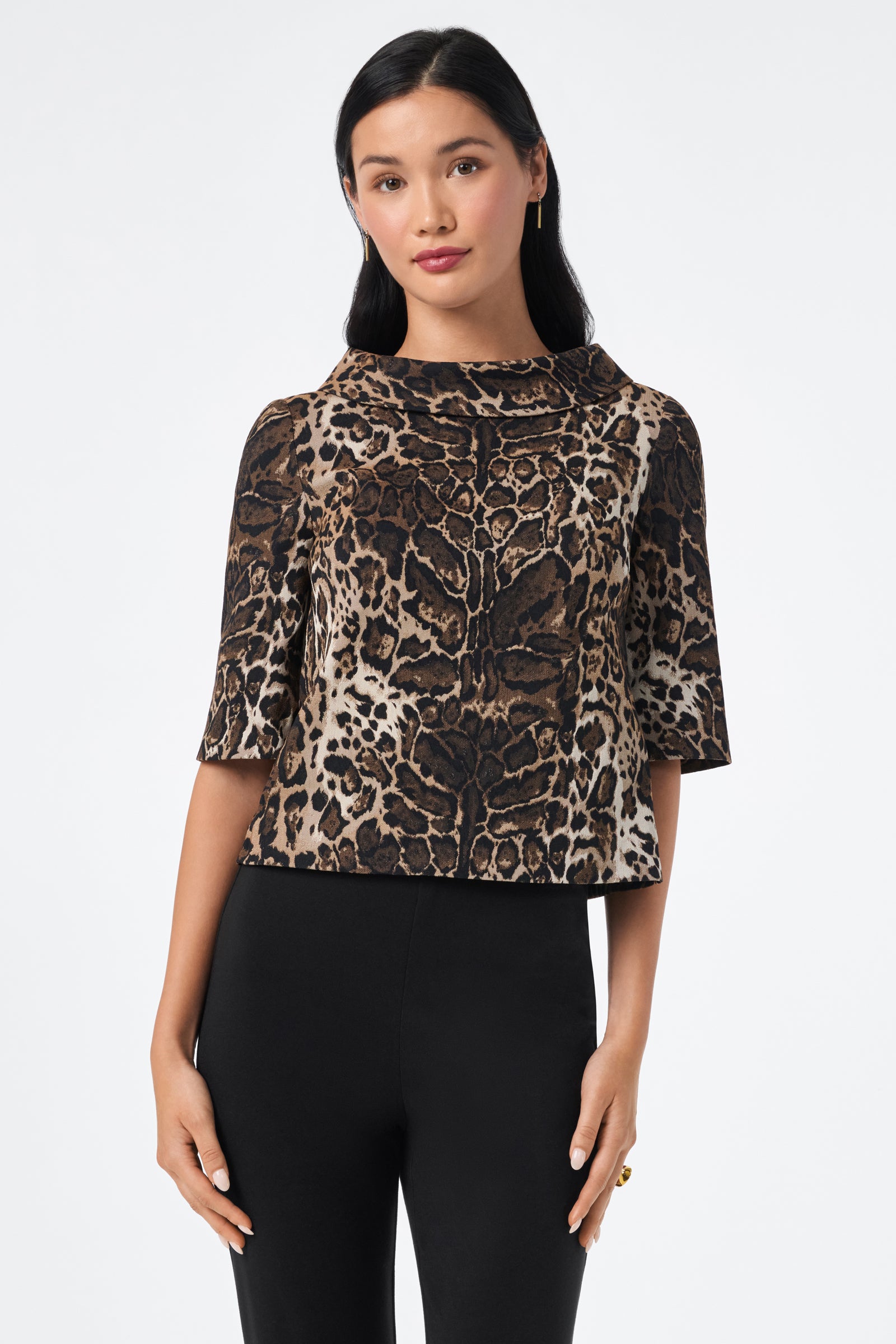 A woman with long black hair wears the Kailee Top, a leopard print short-sleeve style, paired with black pants. She stands against a white background looking at the camera with a neutral expression.