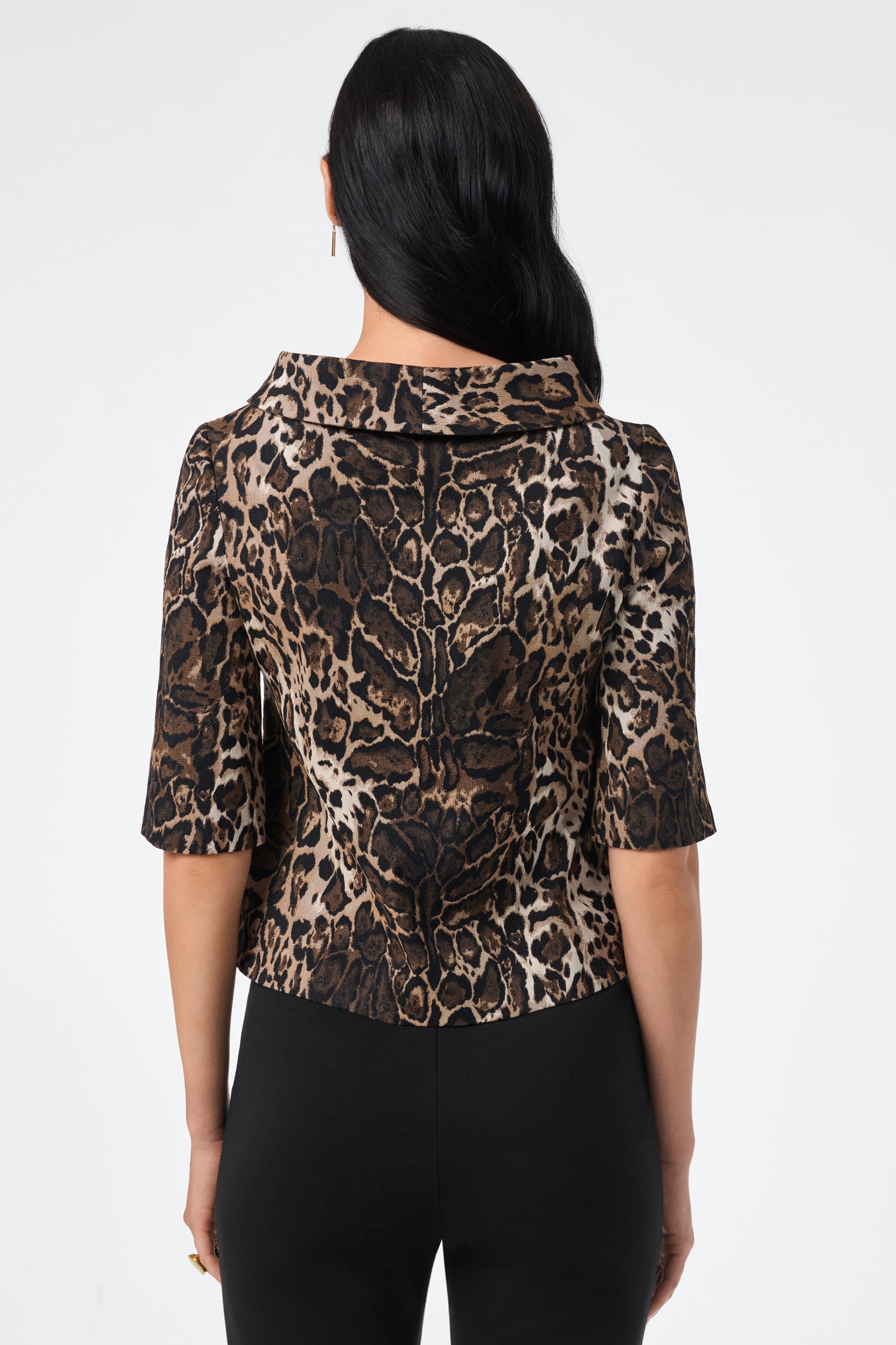 A woman with long dark hair is seen from the back wearing the Kailee Top, a short-sleeve animal-print top with a structured collar, paired with black pants against a plain white background.