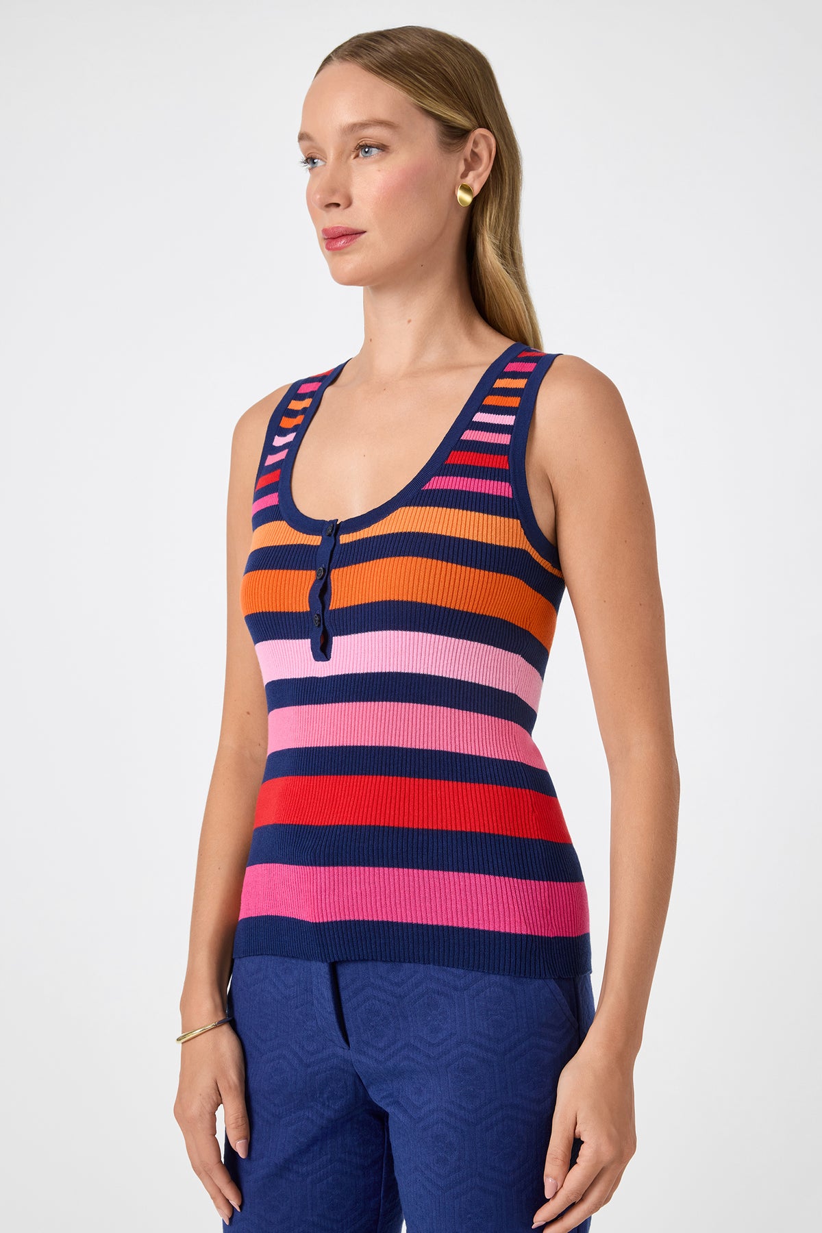 A woman with long light brown hair poses against a plain light background wearing the Arles Scoop Neck Tank‚Äî a sleeveless, knit top with horizontal stripes in navy, orange, red, pink, and purple‚Äîpaired with blue pants.