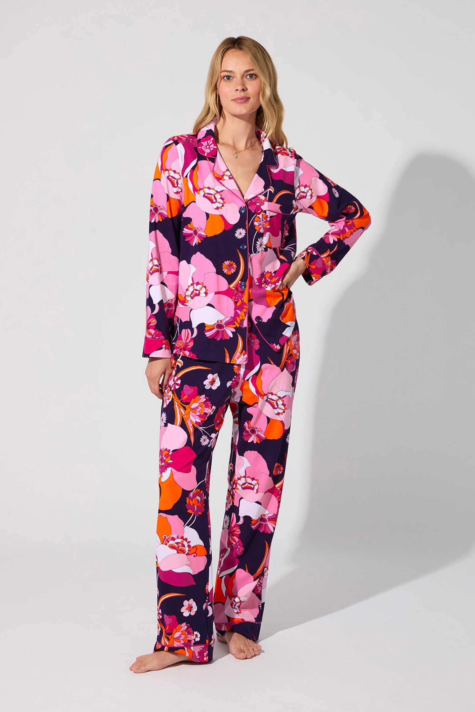 A woman stands barefoot against a plain light background wearing the Floral Medley Classic Pj Set, featuring colorful pink, purple, orange, and white floral patterns. She poses with one hand on her hip.