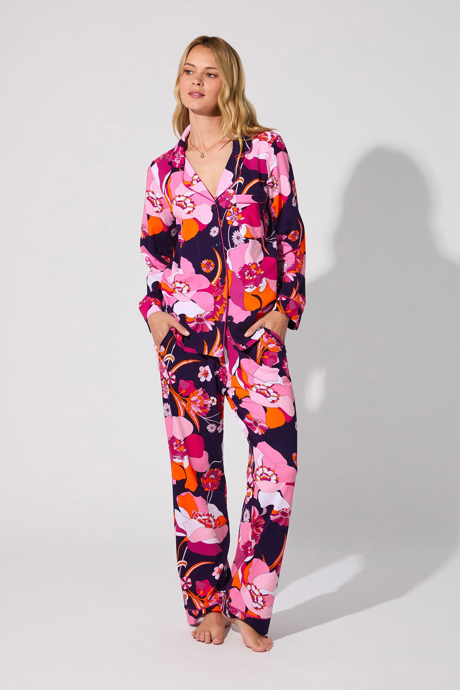 A woman stands barefoot against a plain background, wearing the Floral Medley Classic PJ Set—a matching pajama set with a bold pink, purple, red, and white floral pattern. The top features a collar and buttons; the pants are loose-fitting.