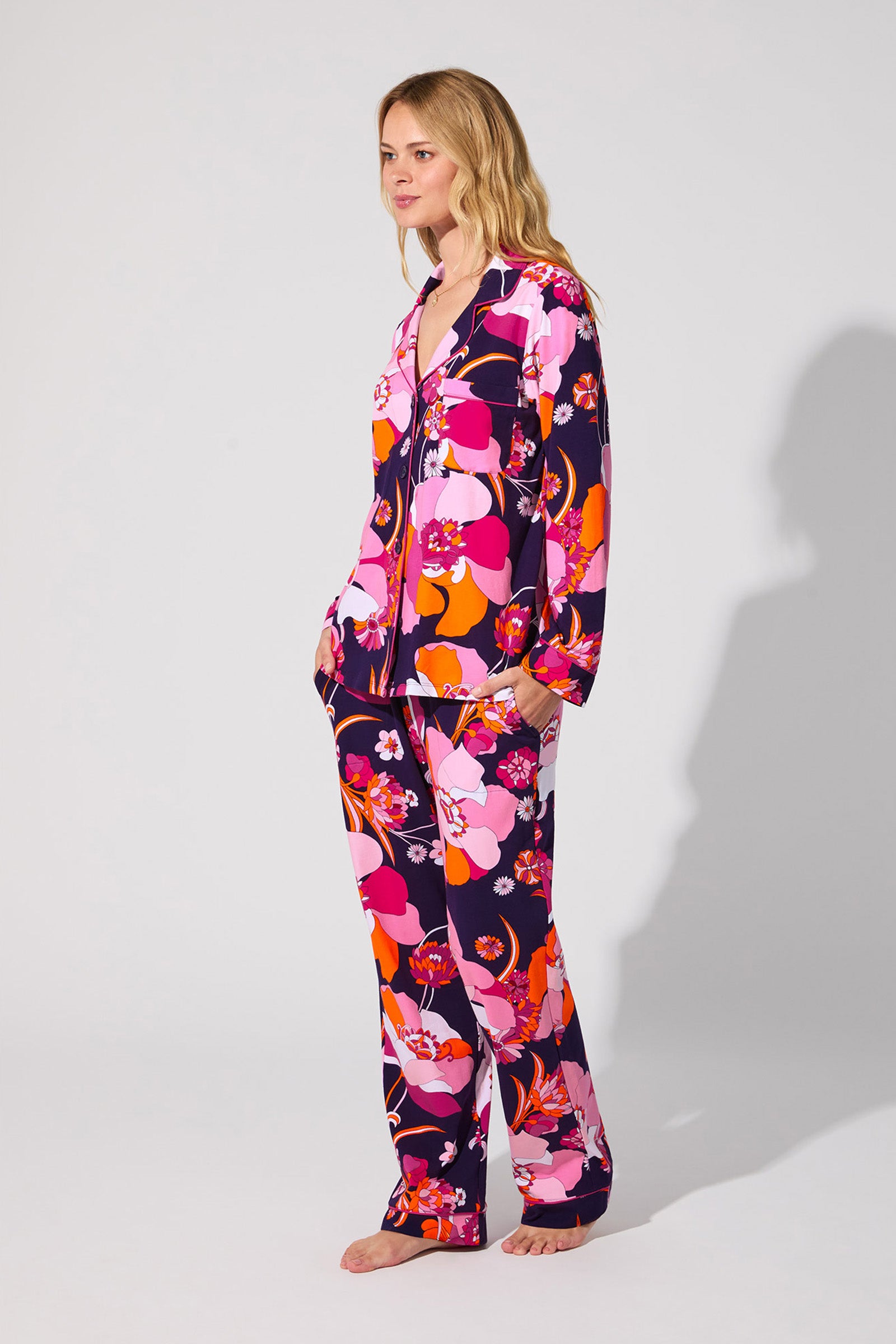 A woman stands barefoot in the Floral Medley Classic Pj Set, featuring a colorful floral pattern in shades of pink, red, orange, and purple, against a plain light gray background.