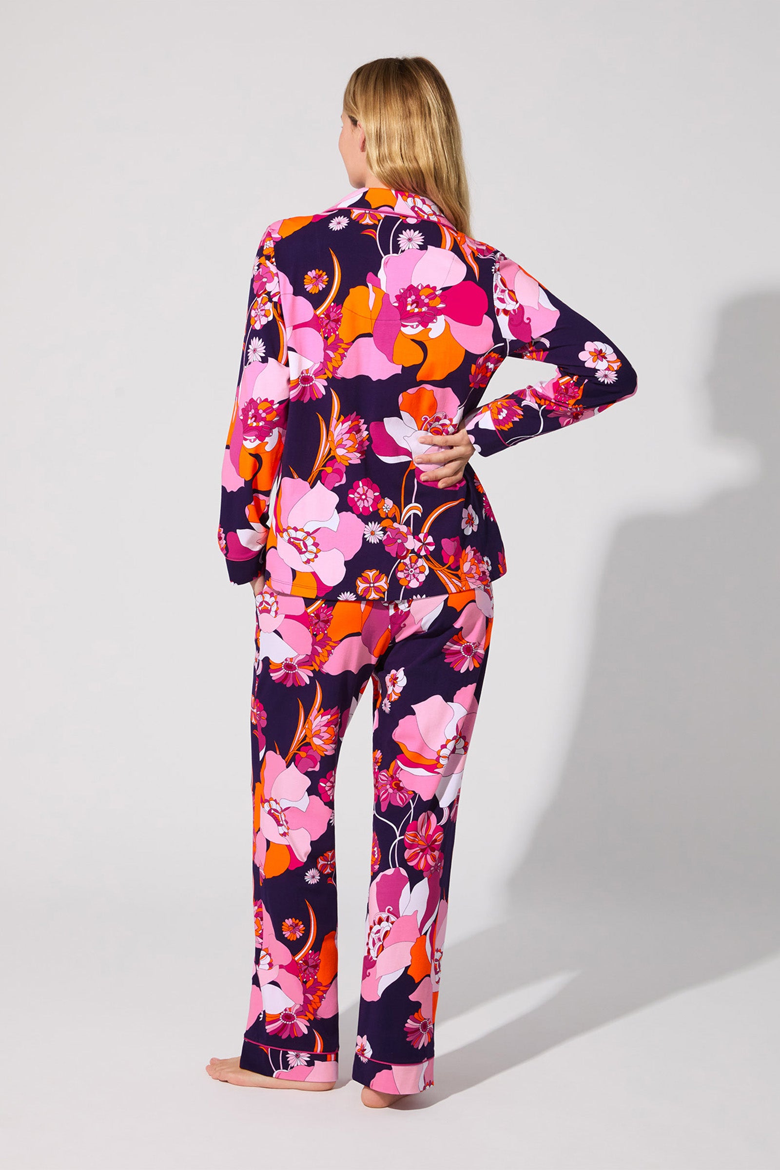 A barefoot person with long blonde hair faces away, wearing the Floral Medley Classic PJ Set—a matching pajama set featuring a bold floral pattern in pink, red, orange, and white on a dark background.