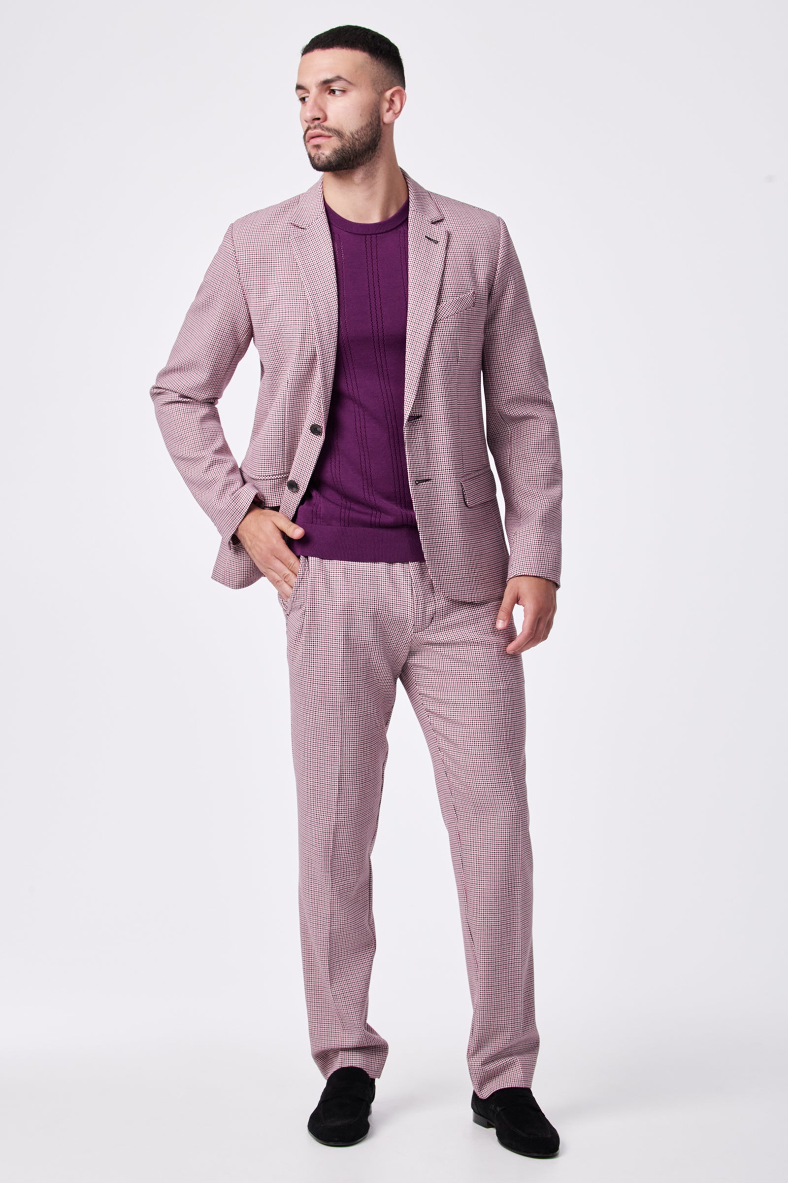A man stands against a white background, looking to the side with one hand in his pocket, wearing the THURSTON BLAZER—a light pink checked suit in viscose, polyester, and elastane—paired with a purple sweater.