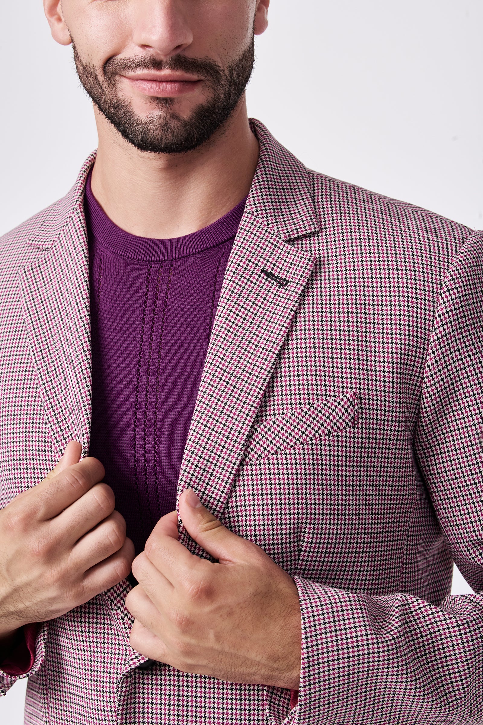 A man wears the THURSTON BLAZER, a pink and white checkered jacket made of polyester and viscose, over a purple knit shirt. Holding the blazer’s lapels, his trimmed beard and slight smile are partially visible.