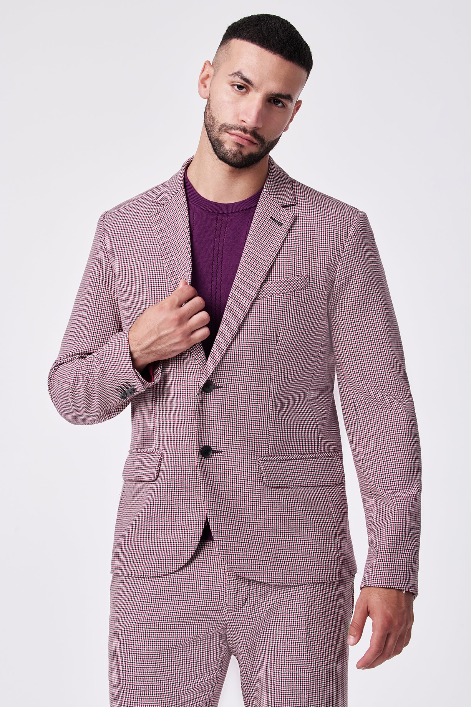 A man with short dark hair and a beard wears the THURSTON BLAZER, a pink checked polyester-viscose suit, over a purple shirt. He stands against a plain white background, holding the jacket lapel and looking at the camera.