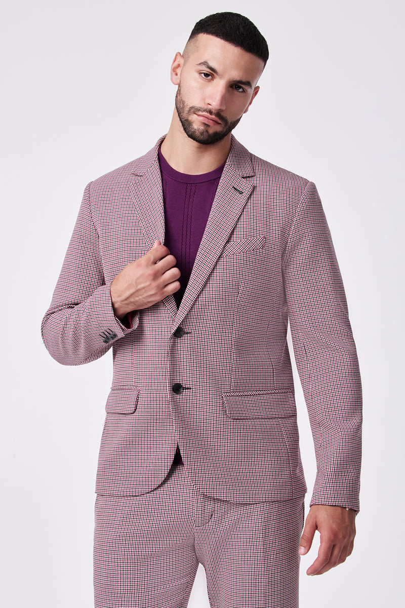 A man with short dark hair and a beard wears the THURSTON BLAZER, a pink checked polyester-viscose suit, over a purple shirt. He stands against a plain white background, holding the jacket lapel and looking at the camera.