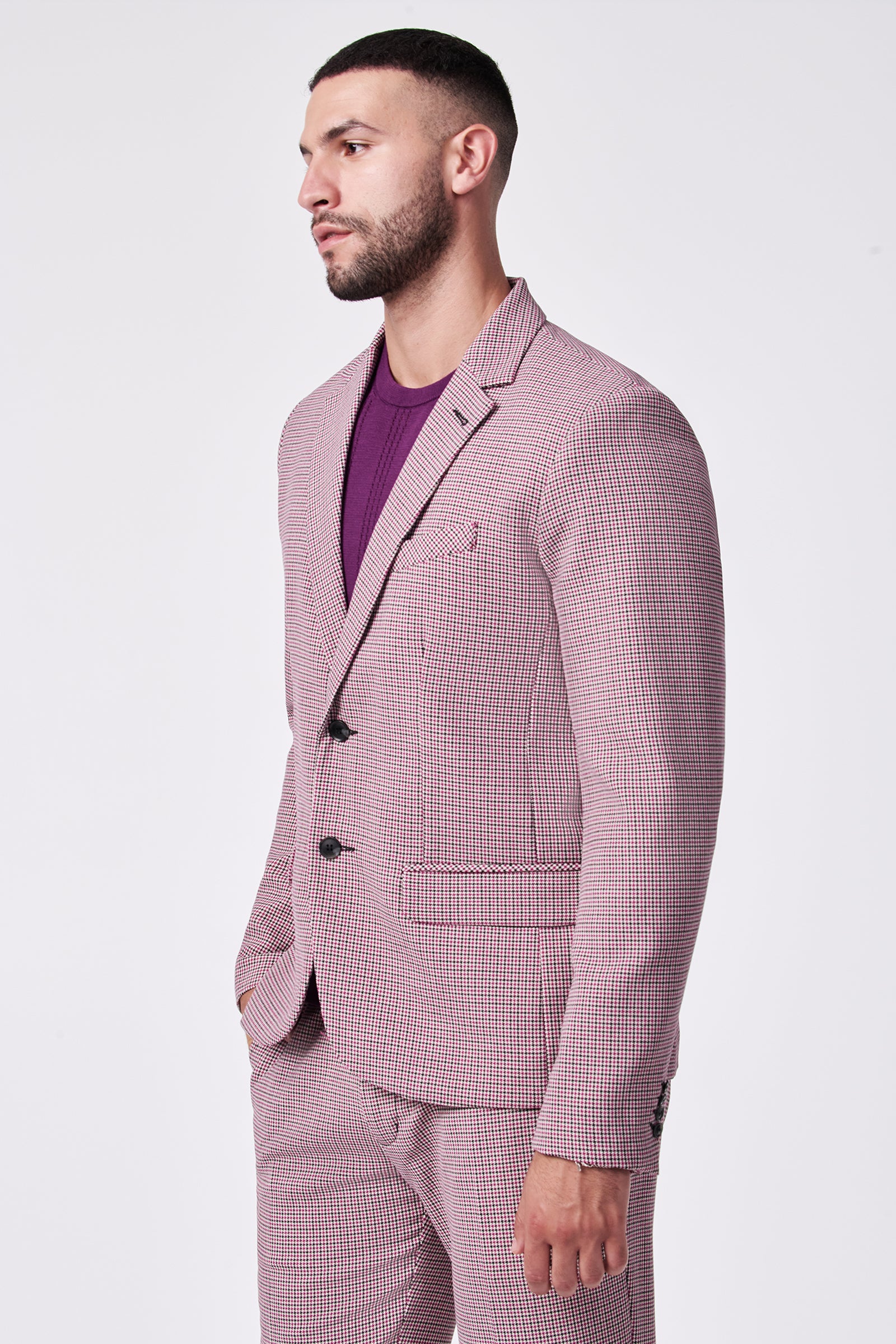 Man with short dark hair and beard wearing the THURSTON BLAZER—a light pink and white checked suit in a soft viscose blend with black buttons—paired with a purple shirt, standing against a plain light background.