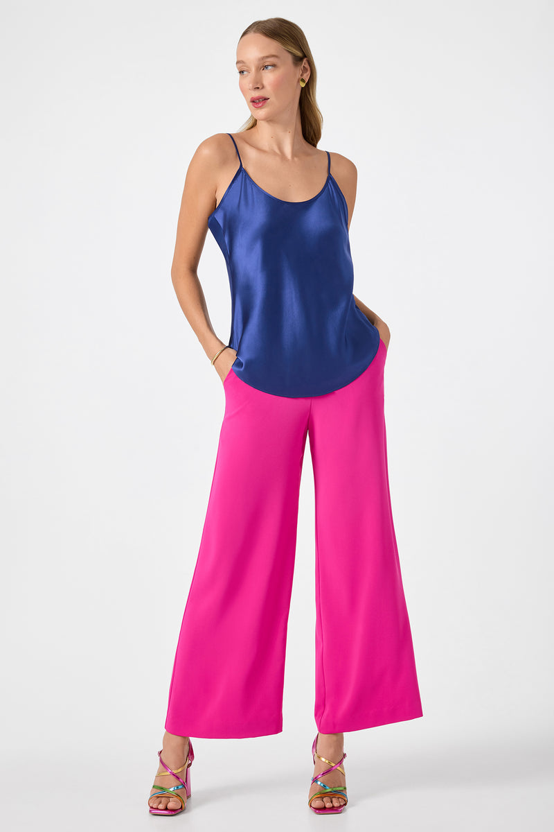 A woman with long hair and a neutral expression stands against a white background, wearing shiny blue camisole, strappy heeled sandals, and bright pink Verity 3 Wide-Leg Pants with her hands in her pockets.